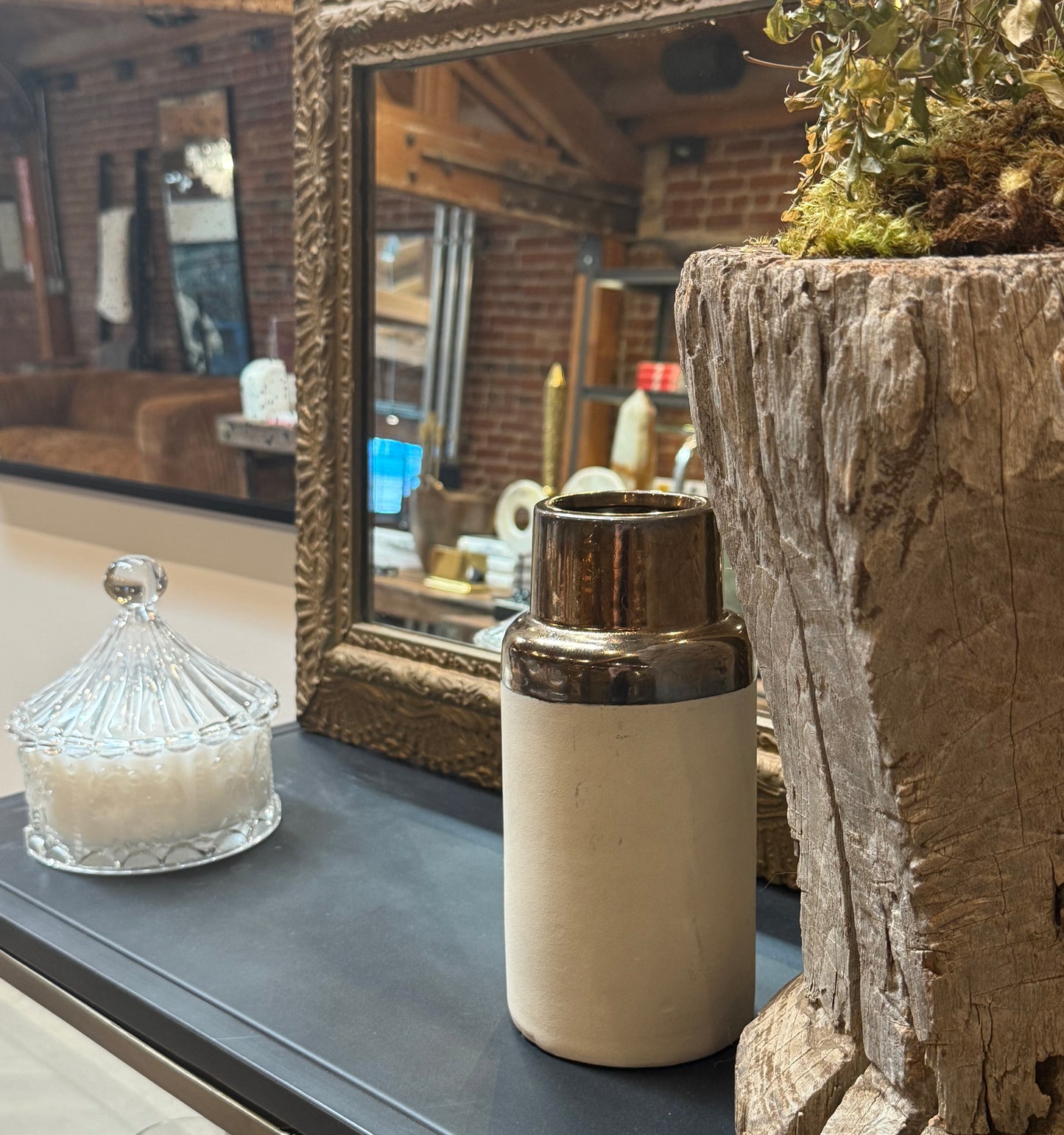 A glass jar, the Metallic and Ceramic Vase (measuring 4.25″ in diameter and 9.25″ high), and a potted plant sit on a black surface in front of a framed mirror, reflecting a cozy room with brick walls and brown furniture.