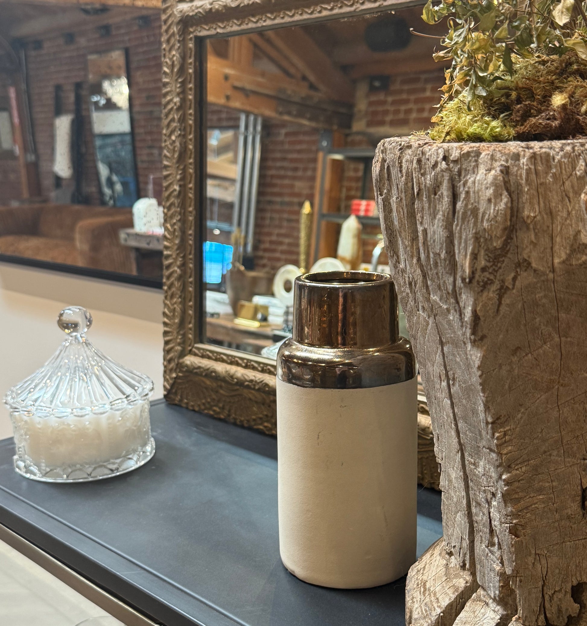 A glass jar, the Metallic and Ceramic Vase (measuring 4.25″ in diameter and 9.25″ high), and a potted plant sit on a black surface in front of a framed mirror, reflecting a cozy room with brick walls and brown furniture.