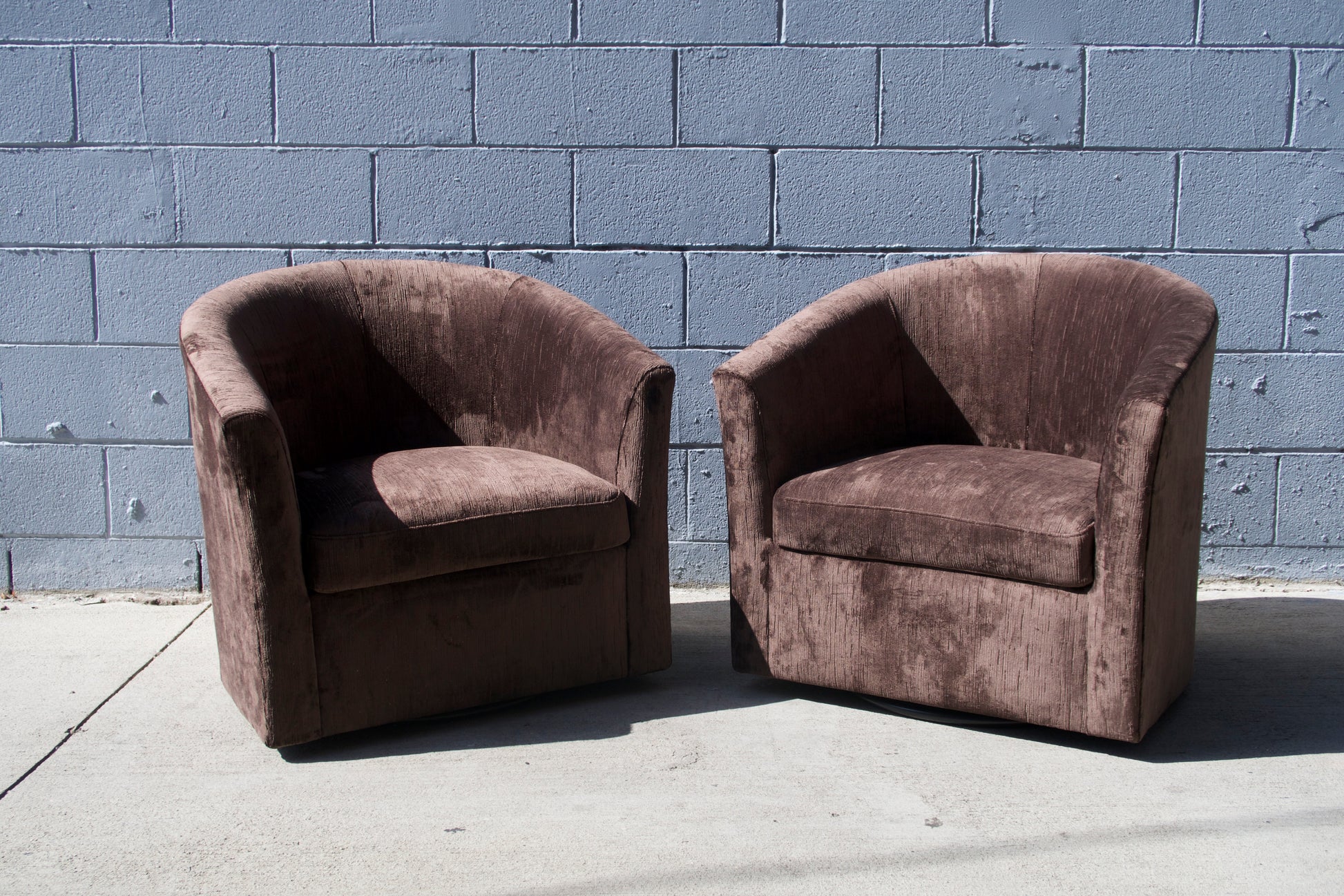 Two Vintage Chocolate Velvet Swivel Chairs, each 29.5"W x 26"D x 28"H, sit side by side on a concrete surface outdoors in front of a gray cinder block wall, illuminated by direct sunlight.
