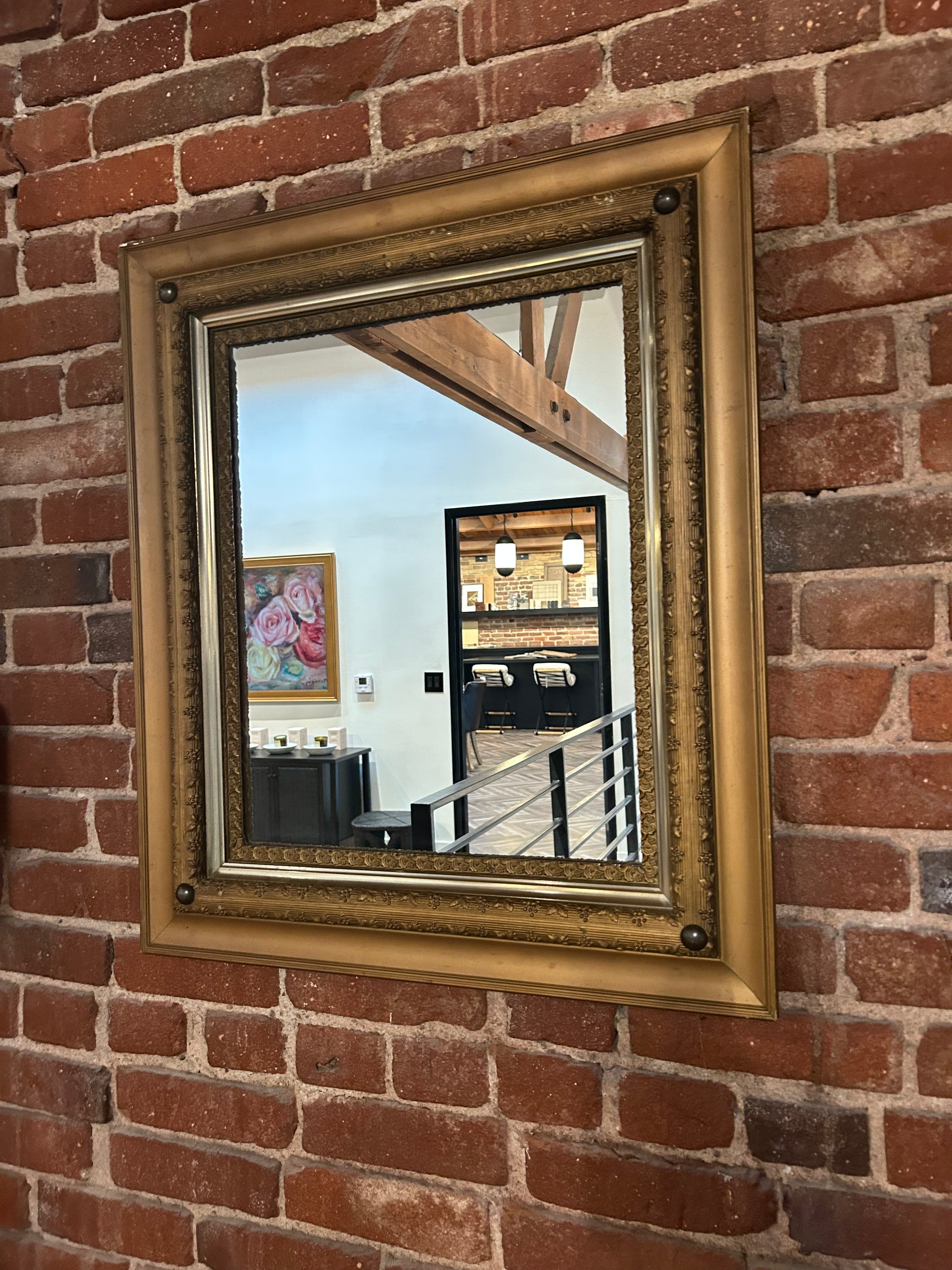 A Vintage Rectangular Gilt Mirror hangs on a red brick wall, reflecting a modern interior with a painting, pendant lights, and part of a staircase railing.