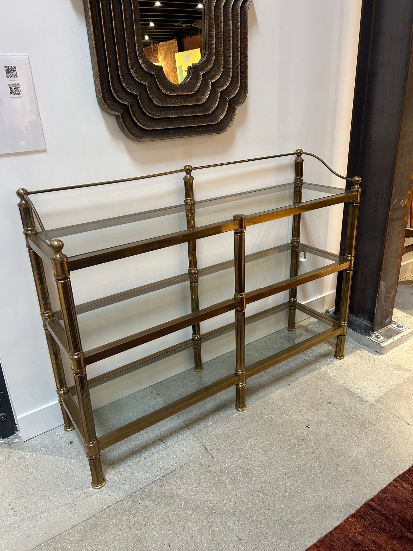 The Vintage Brass & Glass Console, measuring 52"W x 15"D x 39"H with three shelves, stands against a white wall beneath a large wavy-edged brown mirror in a modern showroom.