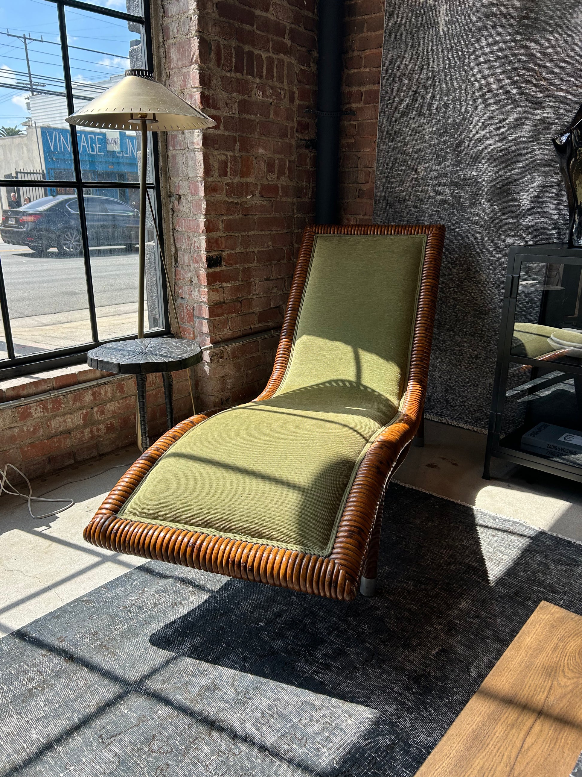 The Vintage Rattan and Green Fabric Chaise, with its curved wicker frame and green upholstery, is ideal for lofts with exposed brick walls, fitting perfectly by a floor lamp on a dark rug near an industrial window.