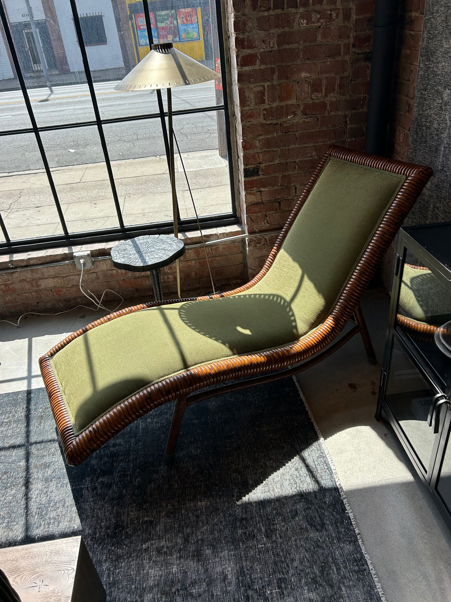 The Vintage Rattan and Green Fabric Chaise sits by a large industrial window, its woven wooden frame and bold hue illuminated by sunlight. Nearby, a round side table and floor lamp accent the exposed brick wall backdrop.