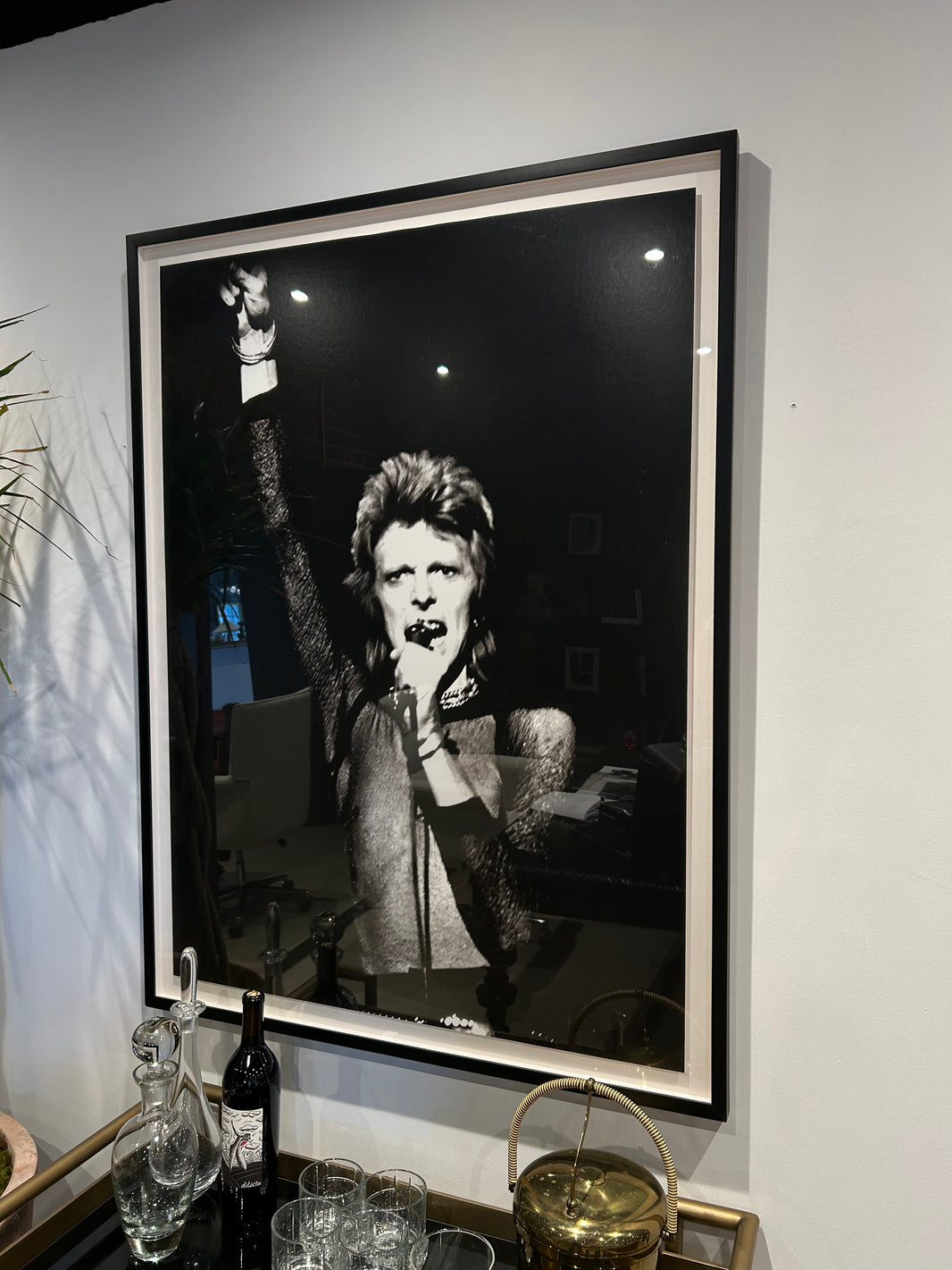 A David Bowie - Framed B/W Gallery Print hangs on a white wall above a bar cart with glassware, a wine bottle, and a gold container; it shows Bowie with spiky hair, holding a microphone and raising one arm.