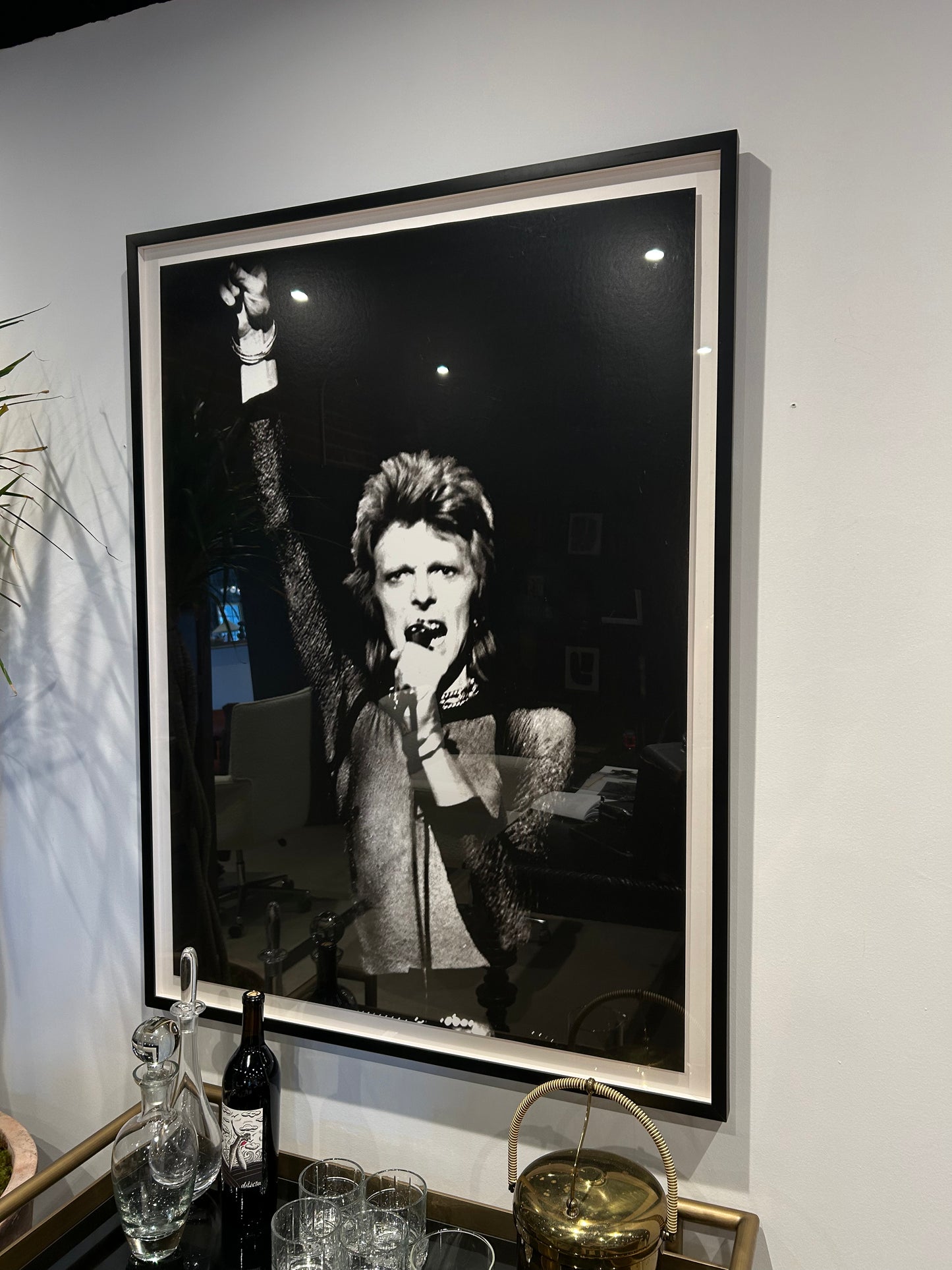 A David Bowie - Framed B/W Gallery Print hangs on a white wall above a bar cart with glassware, a wine bottle, and a gold container; it shows Bowie with spiky hair, holding a microphone and raising one arm.