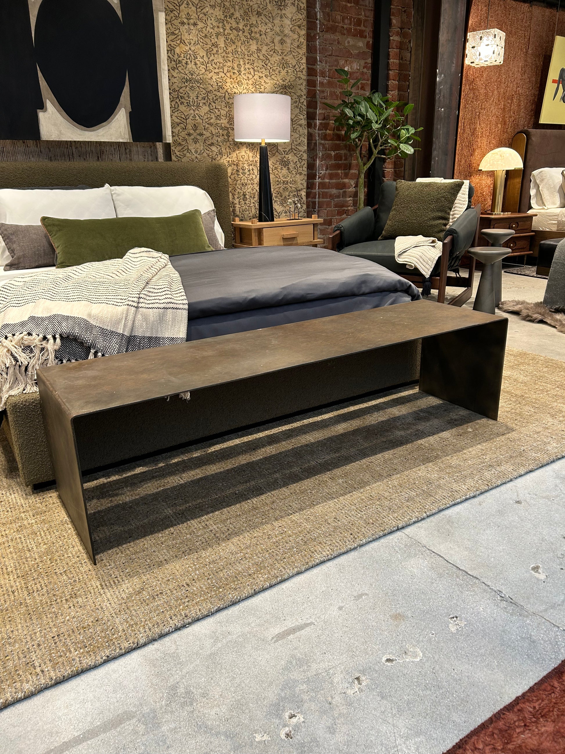 A modern bedroom showcases a Solid Metal Waterfall Bench at the bed’s foot, layered rugs, an upholstered bed, and warm lighting. A sleek media console below a 60-inch TV adds style with cozy blankets and a potted plant.
