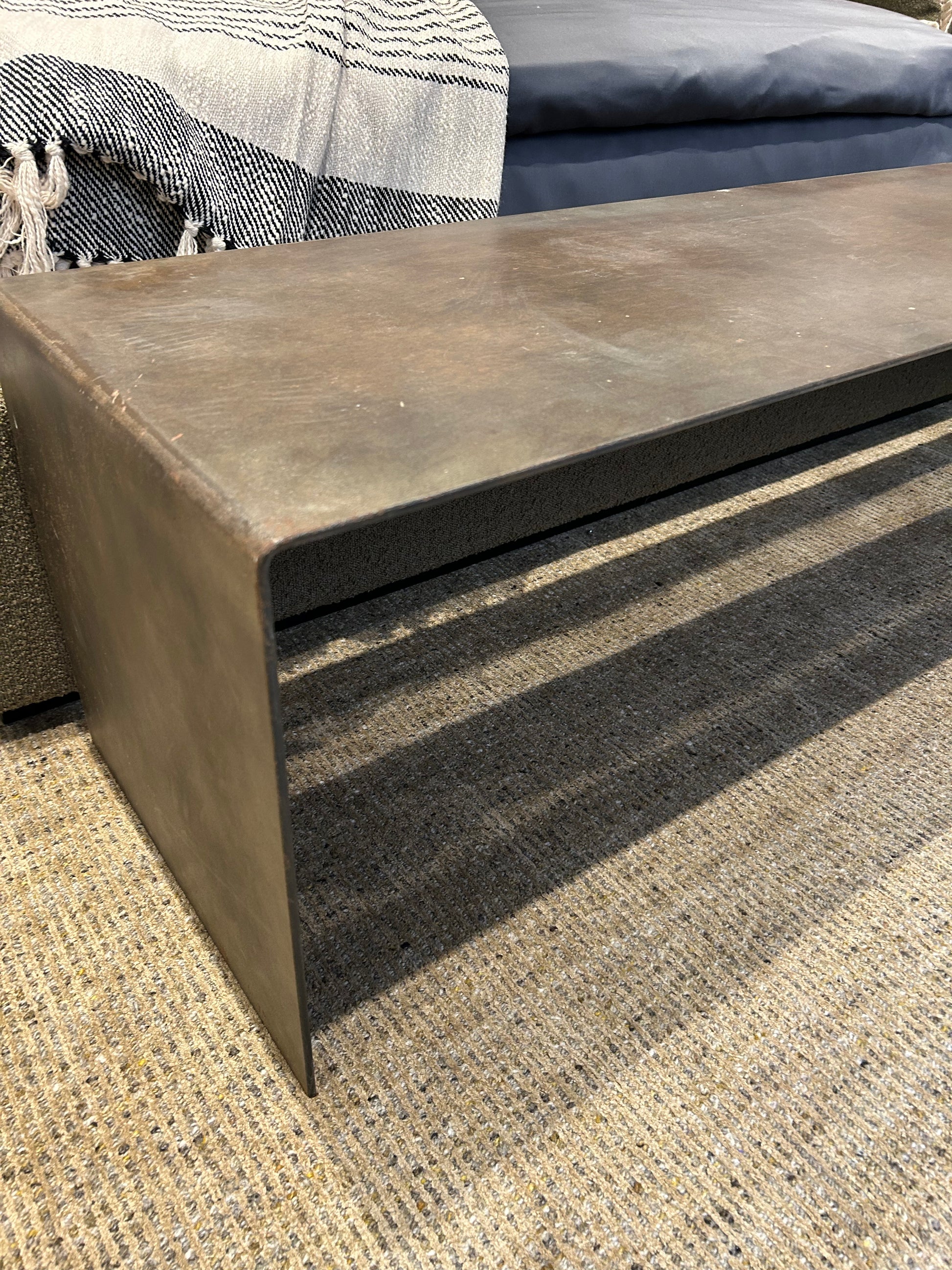 A close-up of the Solid Metal Waterfall Bench with a rust-like finish sits atop a textured tan rug. In the background, a fringed blanket and an elegant modern media console enhance the decor.
