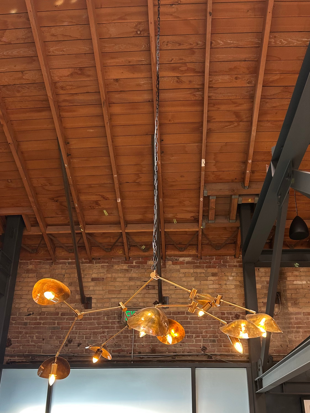The 8-Arm Aged Brass Mantis Chandelier with exposed bulbs hangs from a high wooden ceiling with metal beams, its striking design and dimensions standing out against a brick wall and frosted glass panels in the background.