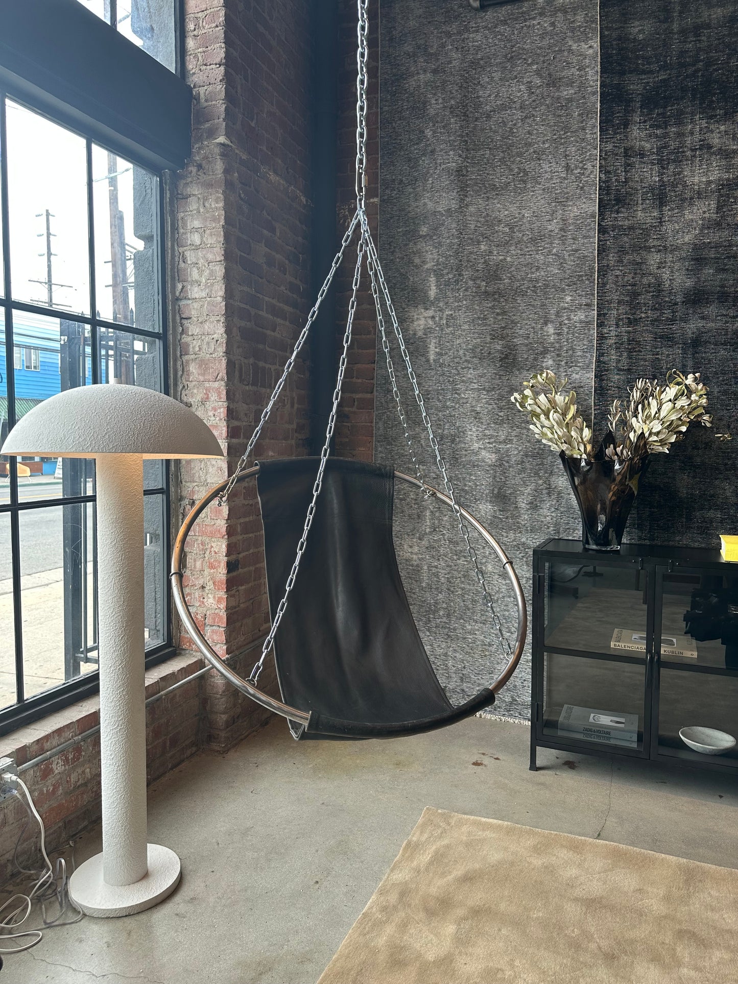 A modern loft corner features a large window, a Vintage Leather Swing Chair with a circular metal frame, a white mushroom floor lamp, a black cabinet, and a vase topped with metallic leaves.