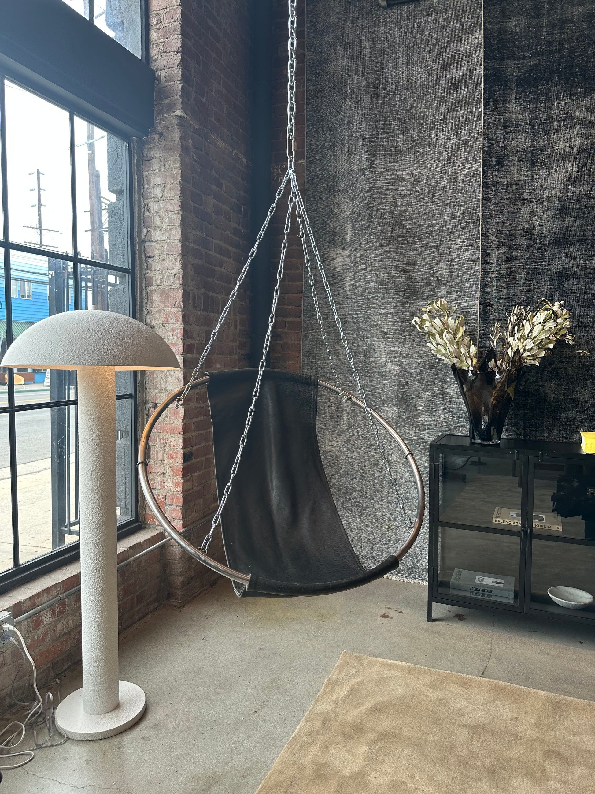 A modern loft corner features a large window, a Vintage Leather Swing Chair with a circular metal frame, a white mushroom floor lamp, a black cabinet, and a vase topped with metallic leaves.