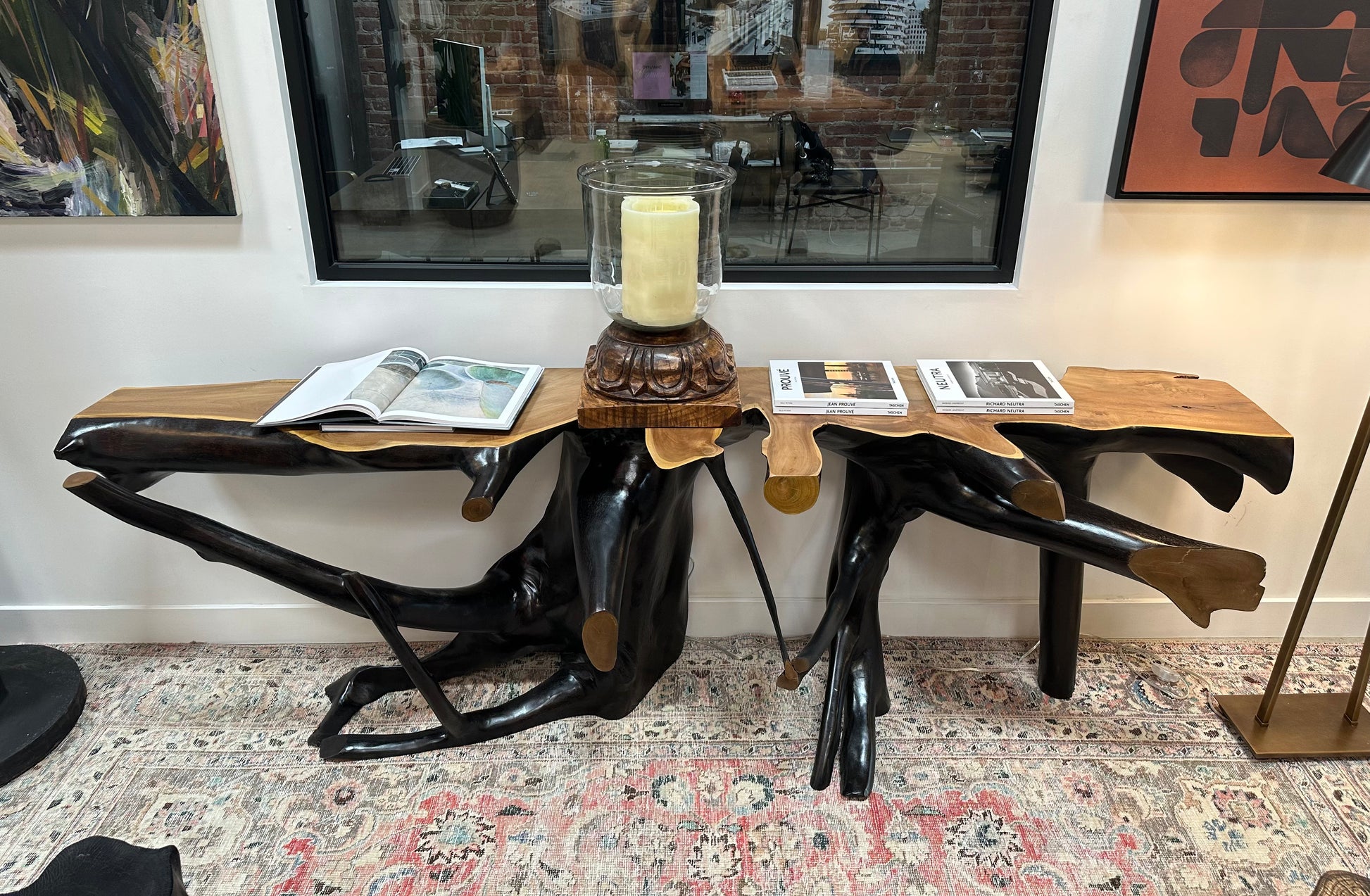 An Organic Ebonized Teak Console Table with branch-like legs displays a large candle and open books next to a modern sofa, set against a window and art-filled wall atop a patterned rug.