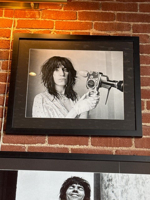 “Patti Smith - Gallery Print on Mat”: Black-and-white portrait of a person with shaggy hair and vintage film camera, wearing a light shirt. Presented in a black frame, 24"H x 28"W, and displayed on a brick wall.
