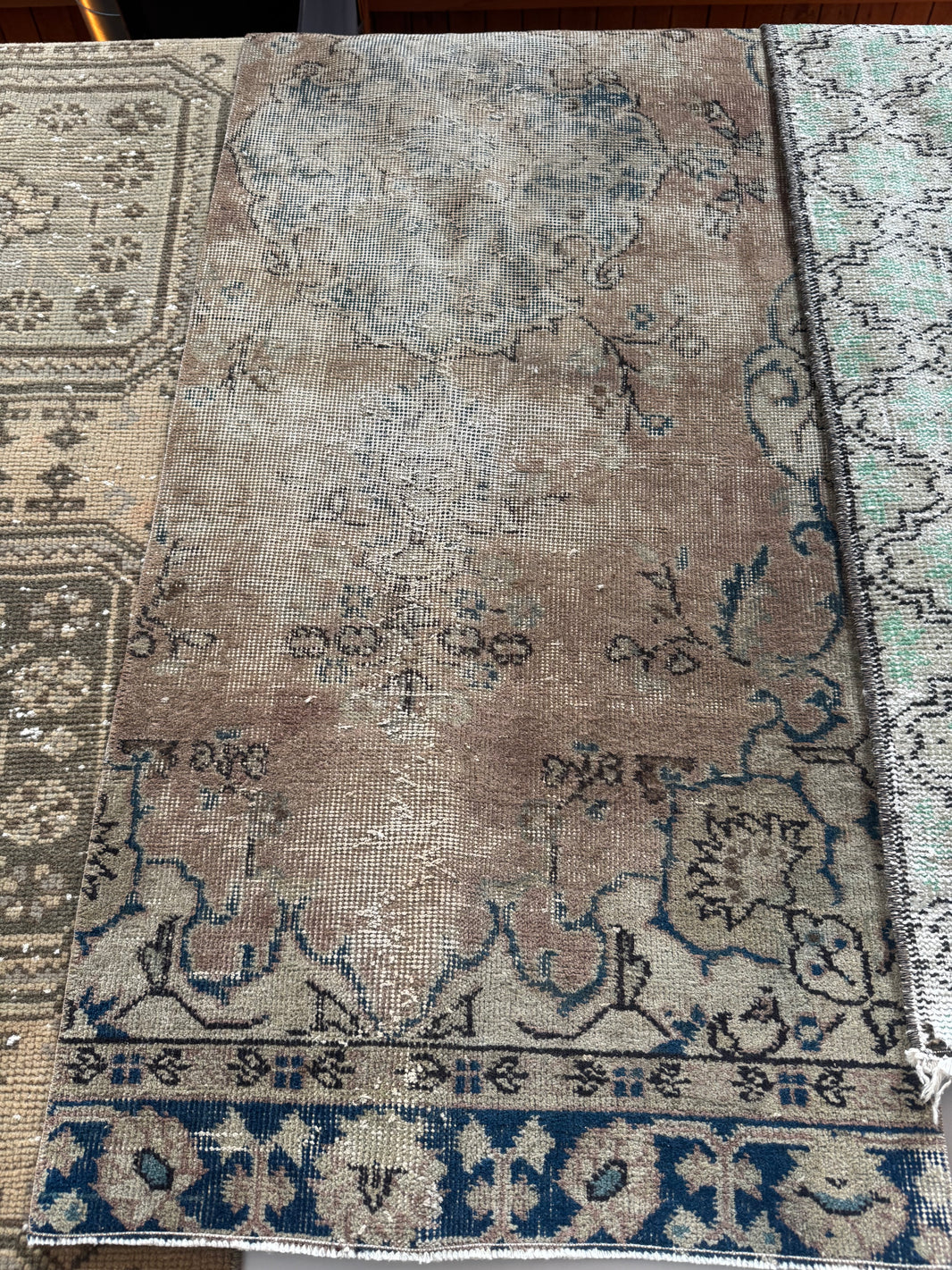 The Vintage Distressed Turkish Runner Rug, in faded brown and blue with muted beige tones and floral geometric patterns, is displayed alongside other patterned rugs.