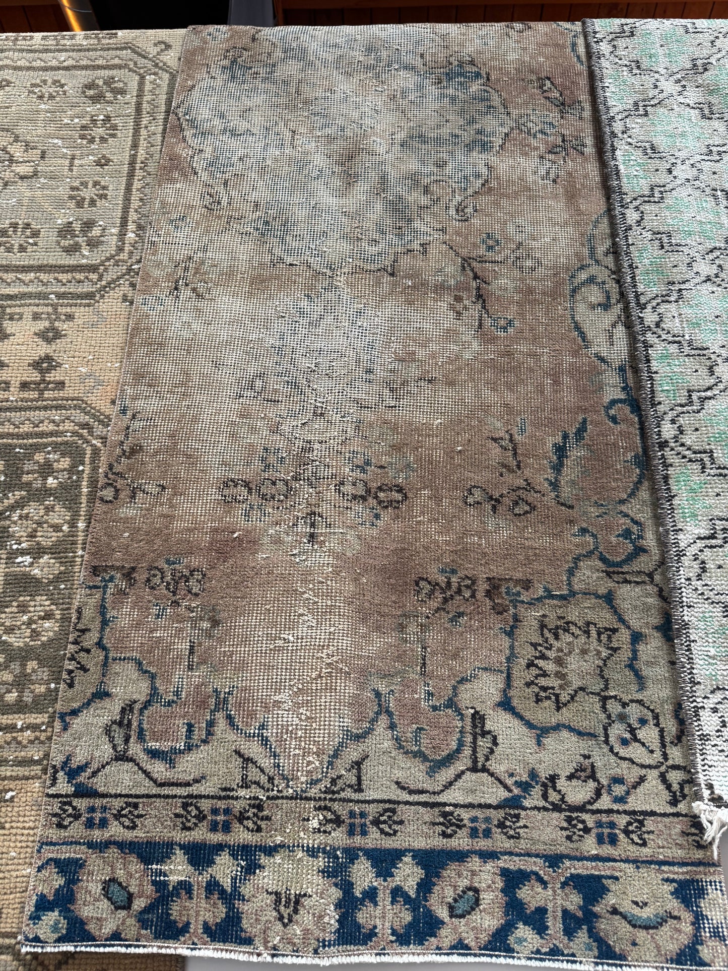 The Vintage Distressed Turkish Runner Rug, in faded brown and blue with muted beige tones and floral geometric patterns, is displayed alongside other patterned rugs.
