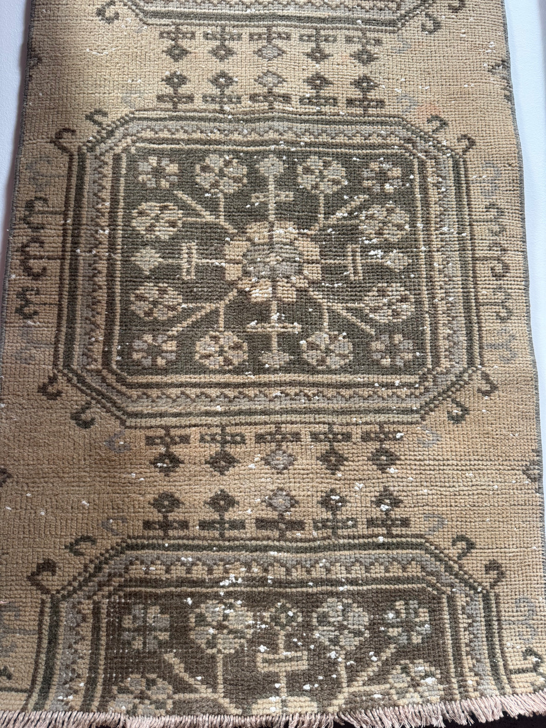 A close-up of a Vintage Turkish Runner Rug (2' x 9') displays worn geometric and floral patterns in muted green on brown, with visible fraying and scattered white specks that highlight its aged, well-used character.