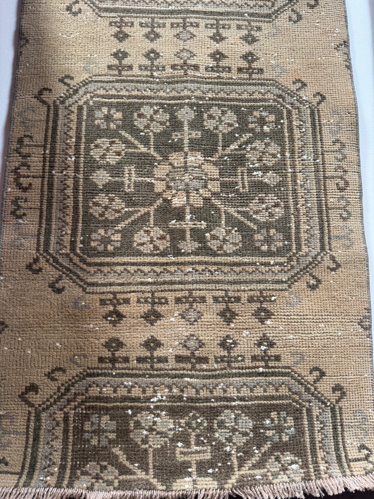 A close-up of a Vintage Turkish Runner Rug (2' x 9') displays worn geometric and floral patterns in muted green on brown, with visible fraying and scattered white specks that highlight its aged, well-used character.