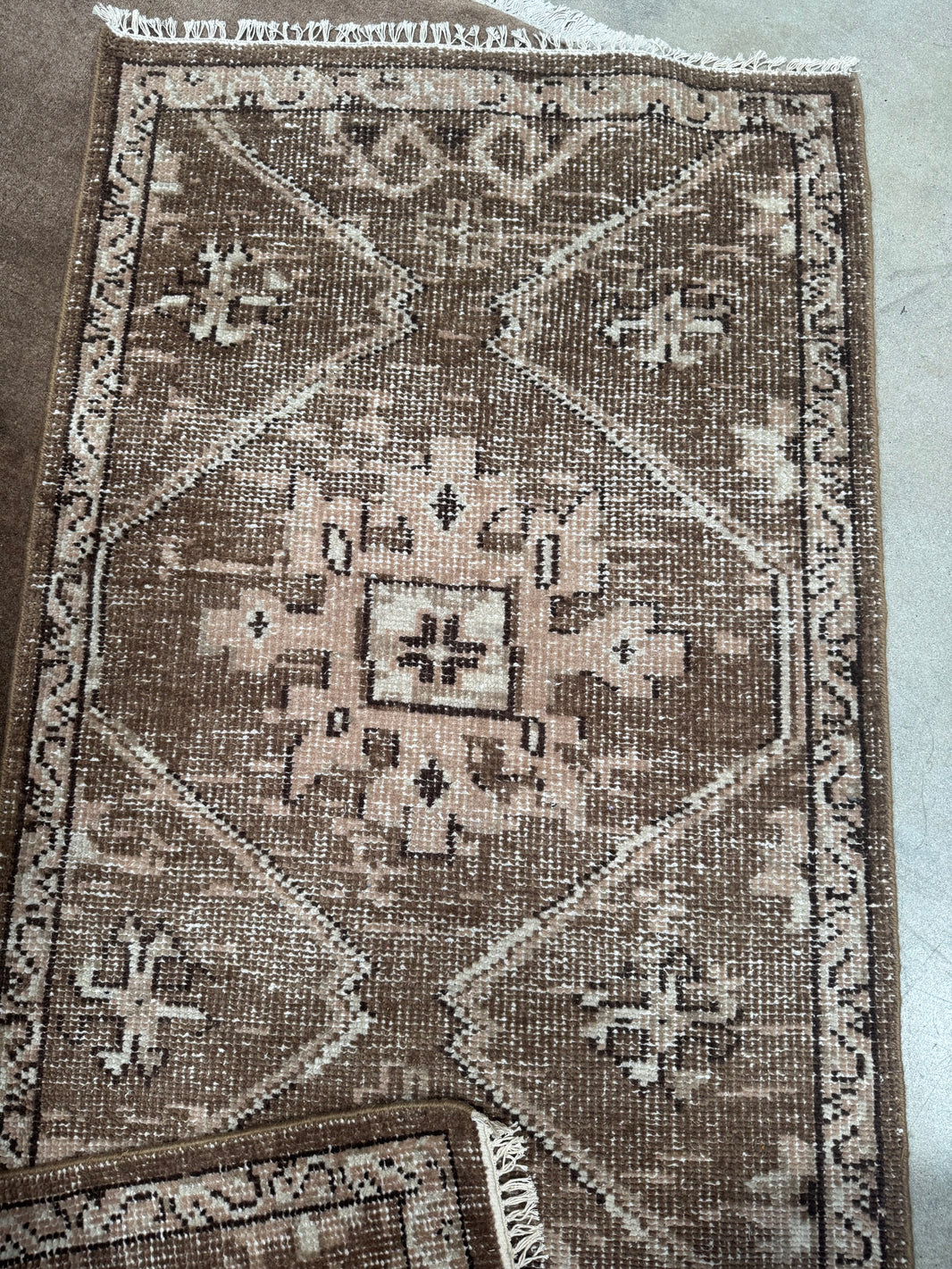 The Vintage Turkish Runner Rug measures 3' x 5', features a brown and beige geometric pattern with central diamond shapes, symmetrical motifs, fringe on one end, and includes a smaller overlapping rug in the corner.