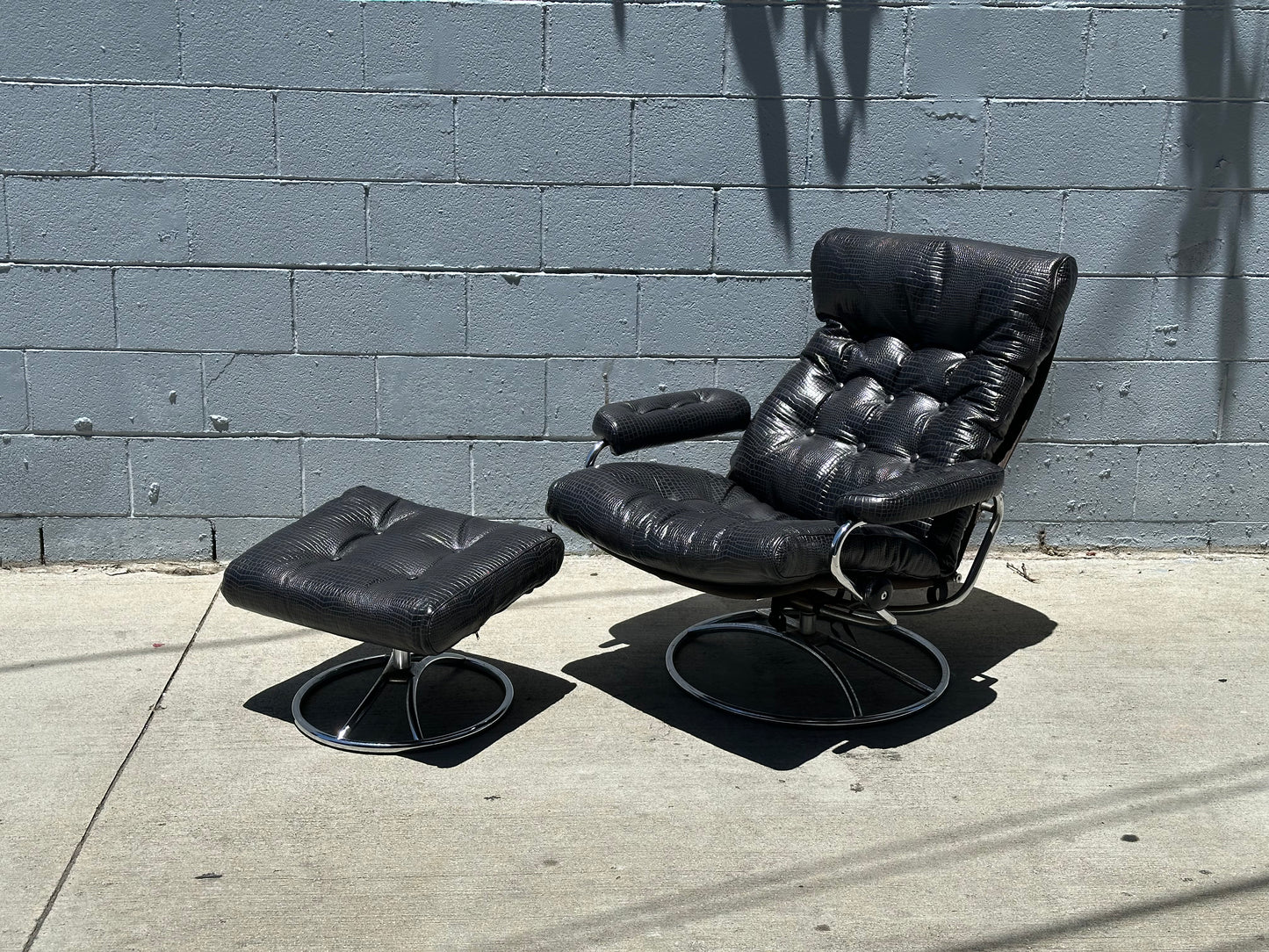 A Vintage Chrome and Leather Recliner & Ottoman set rests on a sunlit concrete sidewalk before a gray brick wall, with tree branch shadows cast across the scene.