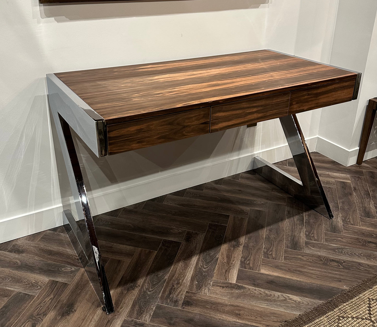 The Vintage Chrome and Walnut Desk features a modern Z shape, three drawers, a polished walnut surface, and angled chrome legs, set against a white wall on a dark herringbone wood floor.