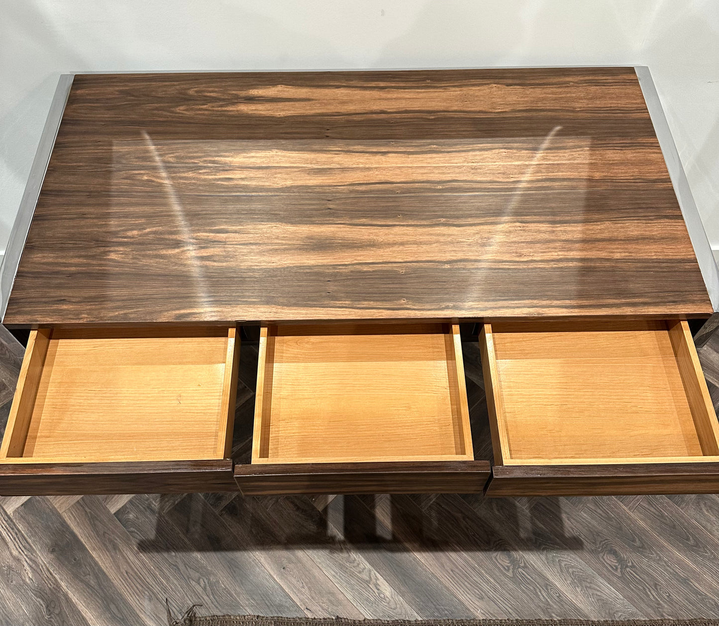 A Vintage Chrome and Walnut Desk with a polished wooden surface and three open empty drawers is viewed from above. This distinctive Z-shaped desk sits on a dark wood floor.