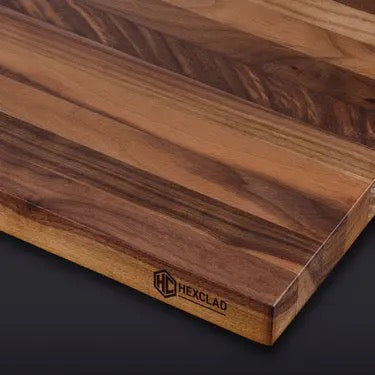 The HexClad Cutting Board is a thick, rectangular board crafted from dark and light wood grains, with the HexClad logo engraved on the side, shown against a dark background.