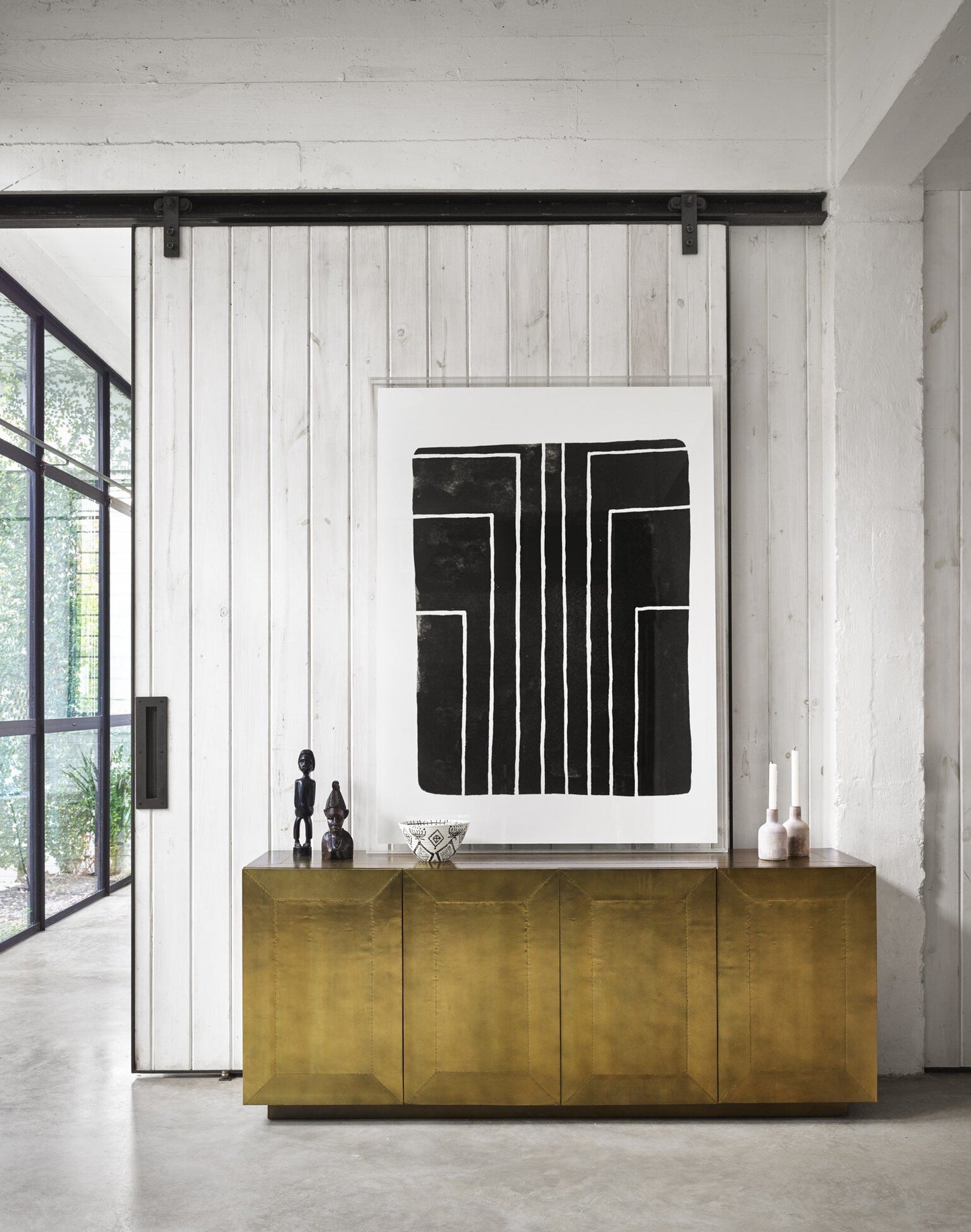 A modern interior features a Brushed Brass Sideboard against a white wood-paneled wall, topped with decorative objects and candles. Above it hangs large abstract black and white art, while glass doors fill the space with natural light.
