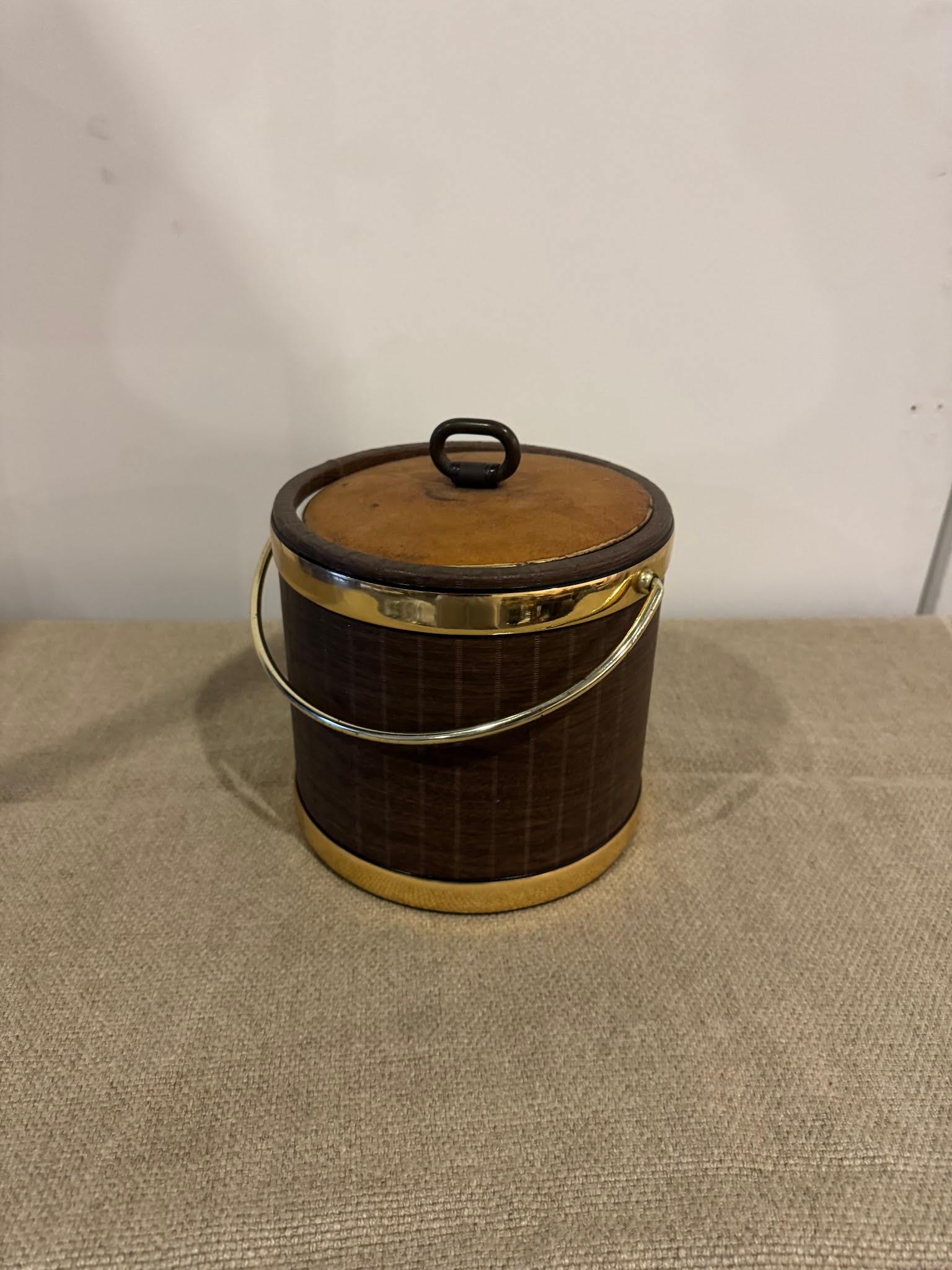 The Ice Bucket, 8.8 inches round in brown with a lid, gold accents, and a handle, rests on a beige textured surface against a plain white wall.