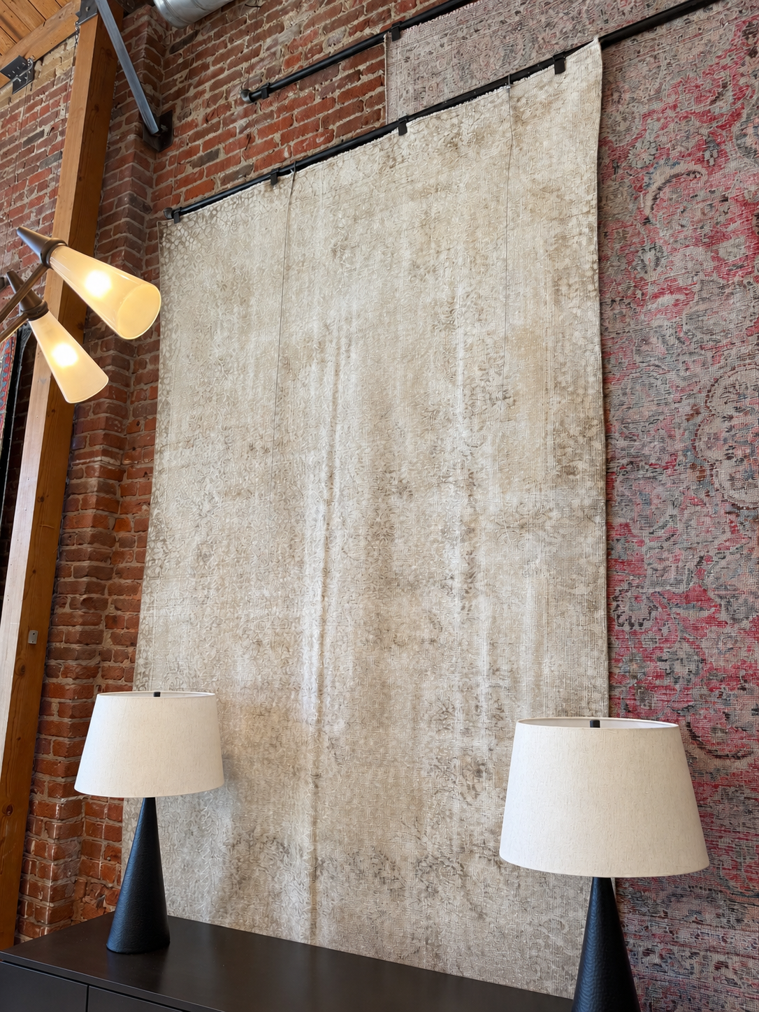 A 6′ x 10′ rectangular Vintage Cream Distressed Wool Area Rug with a faded, distressed pattern hanging on a brick wall between two lamps.
