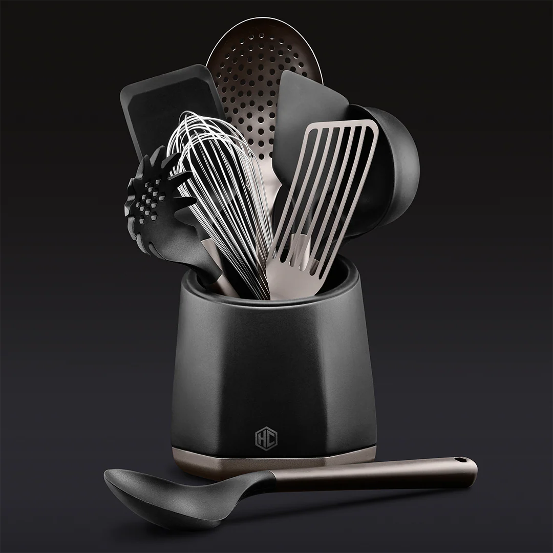 The Hexclad Kitchen Utensil Set, featuring black and silver spatulas, a whisk, and a ladle arranged upright in a matching black holder with a ladle in front, stands out against a dark background.