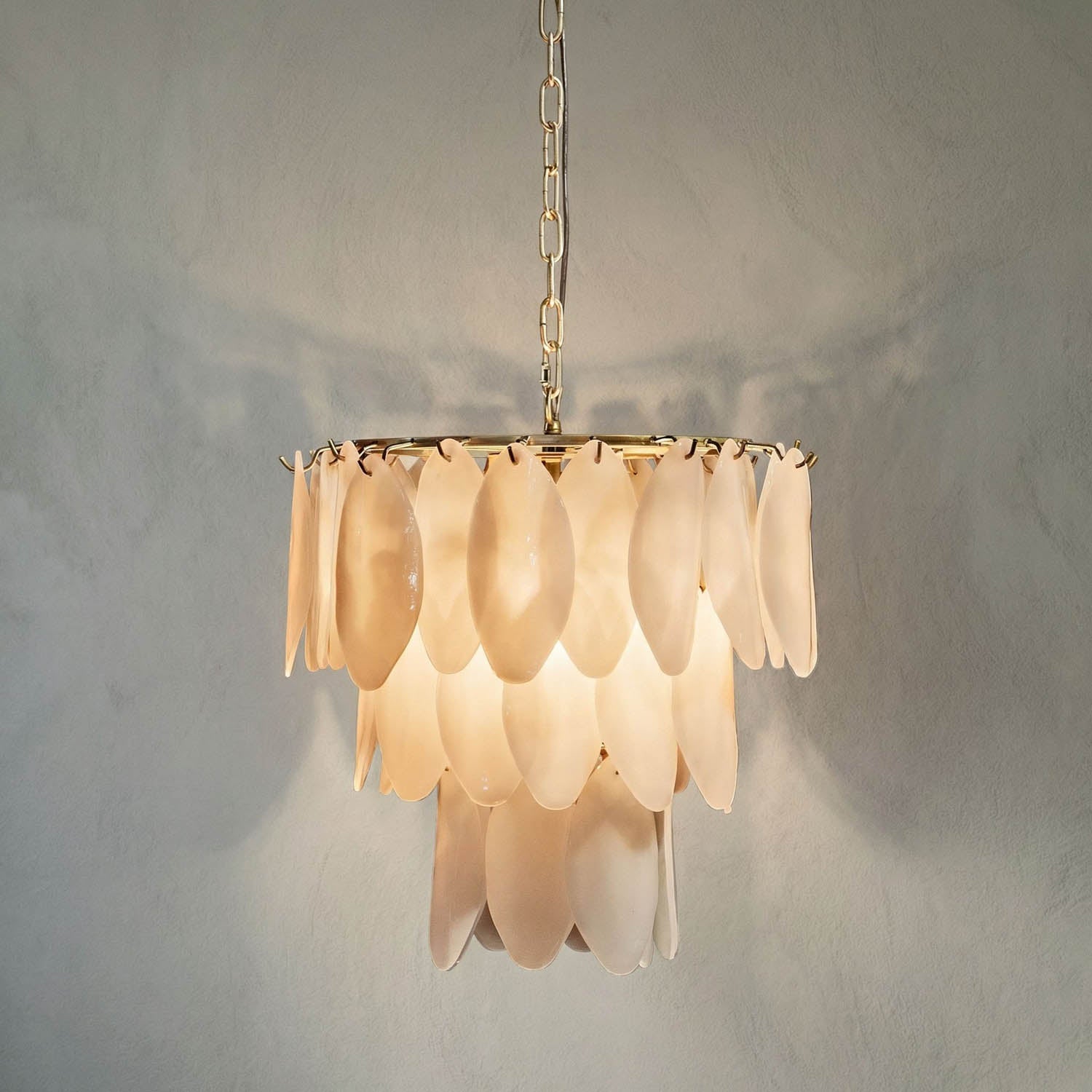 The Opaque Glass Petal Chandelier features overlapping frosted glass petals and hangs from a gold chain, casting a warm, soft glow against light-colored walls.