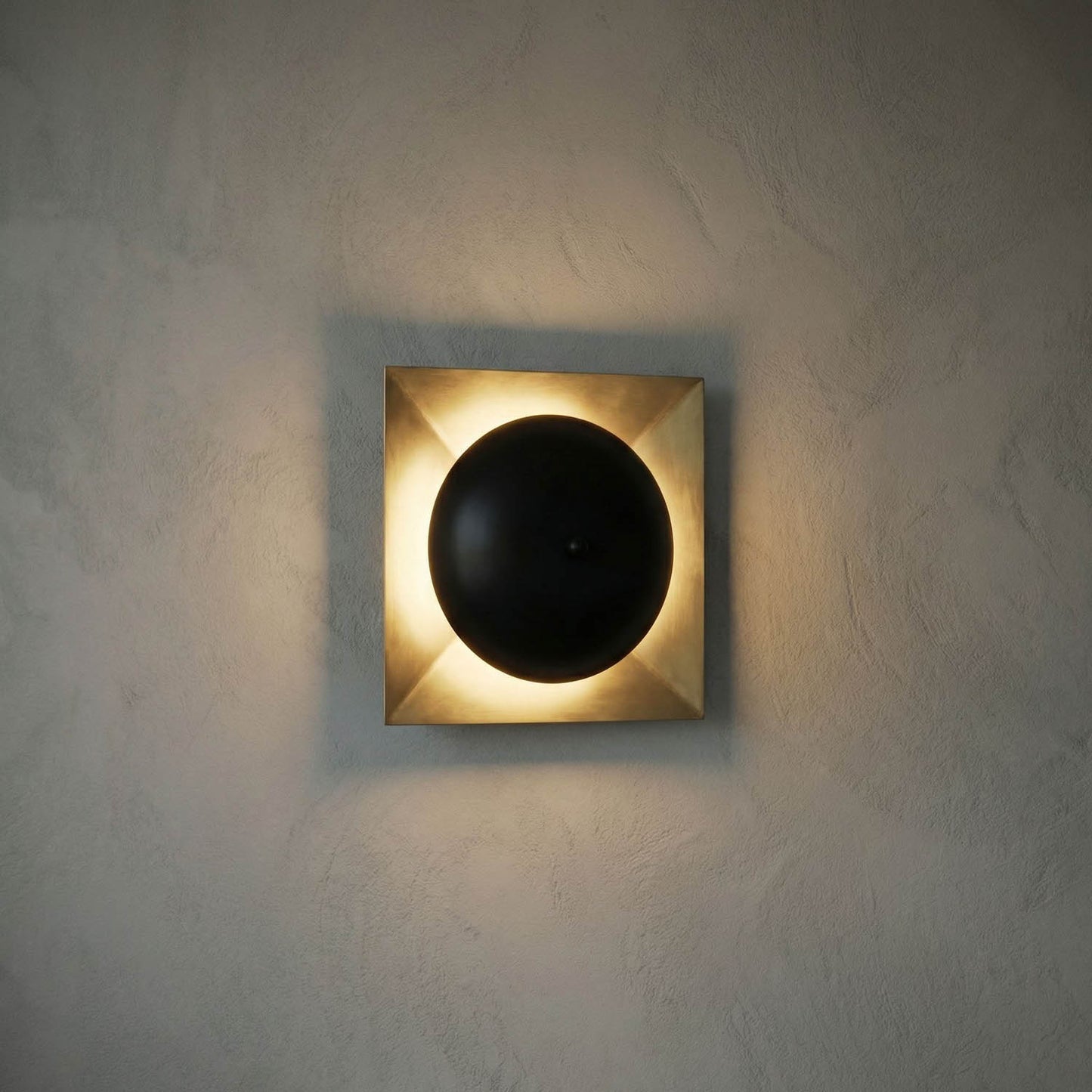 The Solar Eclipse Sconce features a square metallic frame and a central black circle, emitting ambient light that softly glows around the edges, mounted on a textured light wall.