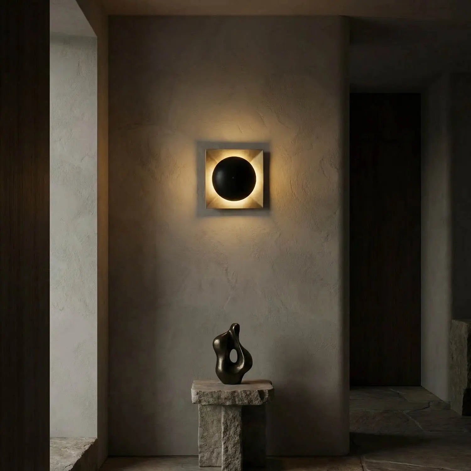 A minimalist room with textured walls features the Solar Eclipse Sconce glowing around a circular center. Below, an abstract dark sculpture rests on a rough stone pedestal, creating a calm, artistic ambiance.