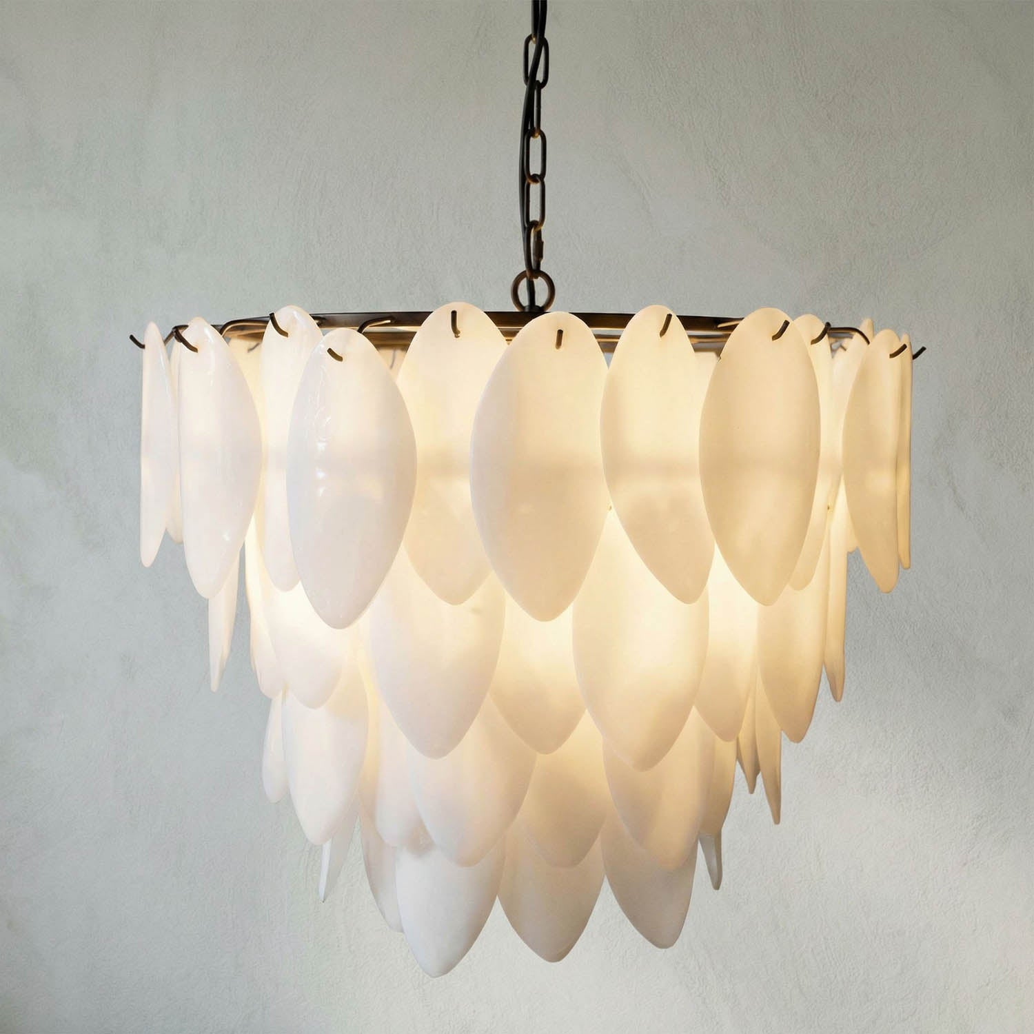 The Opaque Glass Petal Chandelier features layered, oval-shaped opaque glass pieces on a black iron frame, hanging in tiers and beautifully illuminated against a textured wall.
