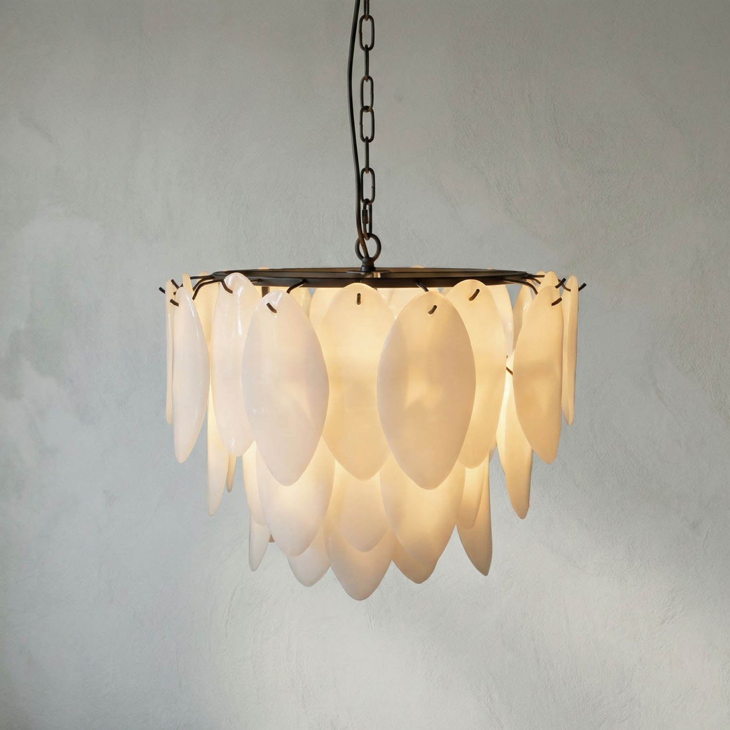 The Opaque Glass Petal Chandelier features layered, oval-shaped translucent panels on a metal frame and chain, creating a soft, glowing effect and adding elegance against a neutral wall.