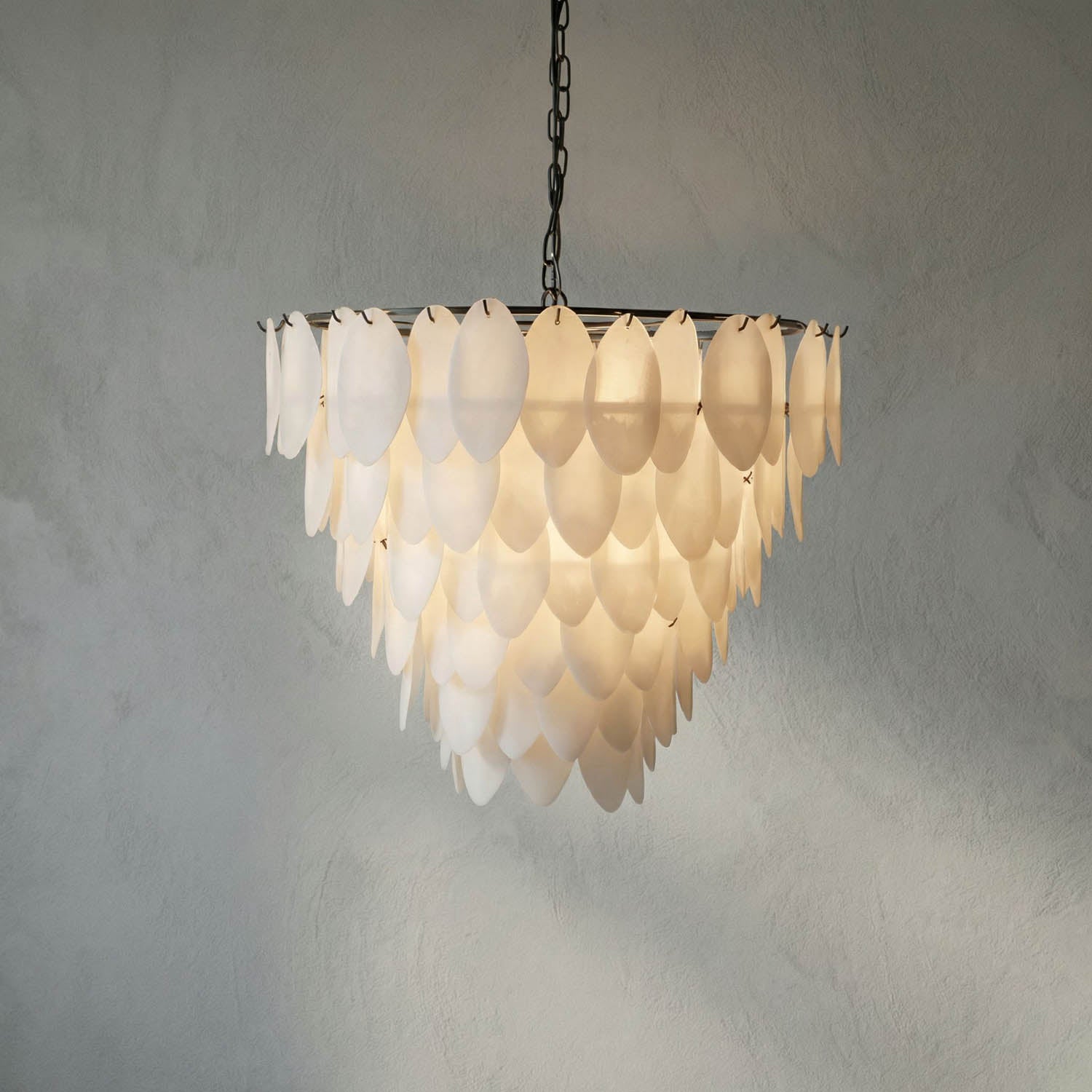 The Opaque Glass Petal Chandelier hangs from the ceiling with layers of translucent, petal-shaped glass that gently diffuse light, offering a modern and elegant touch against a textured gray wall.
