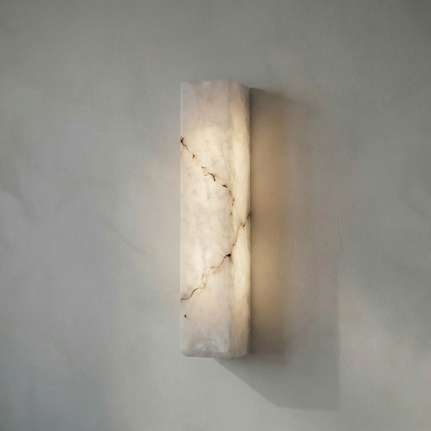 The Rectangular Sconce is a wall-mounted light made of white marble with subtle brown veining, casting a soft glow onto smooth, light-colored walls for a contemporary look.