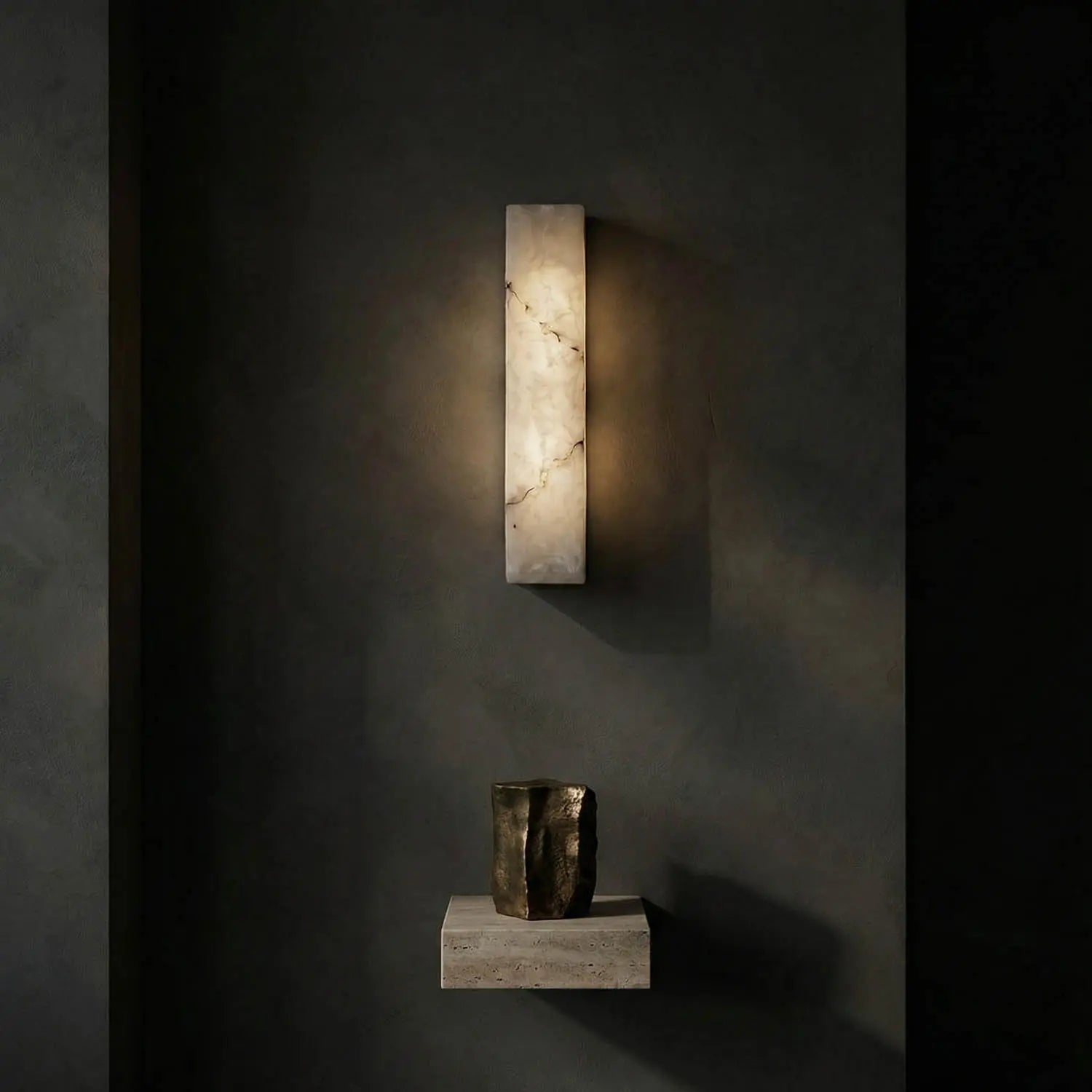 The Rectangular Sconce casts a warm glow on a dark wall while a small stone shelf beneath supports an abstract metallic vase, creating an elegant, minimalist contemporary look.