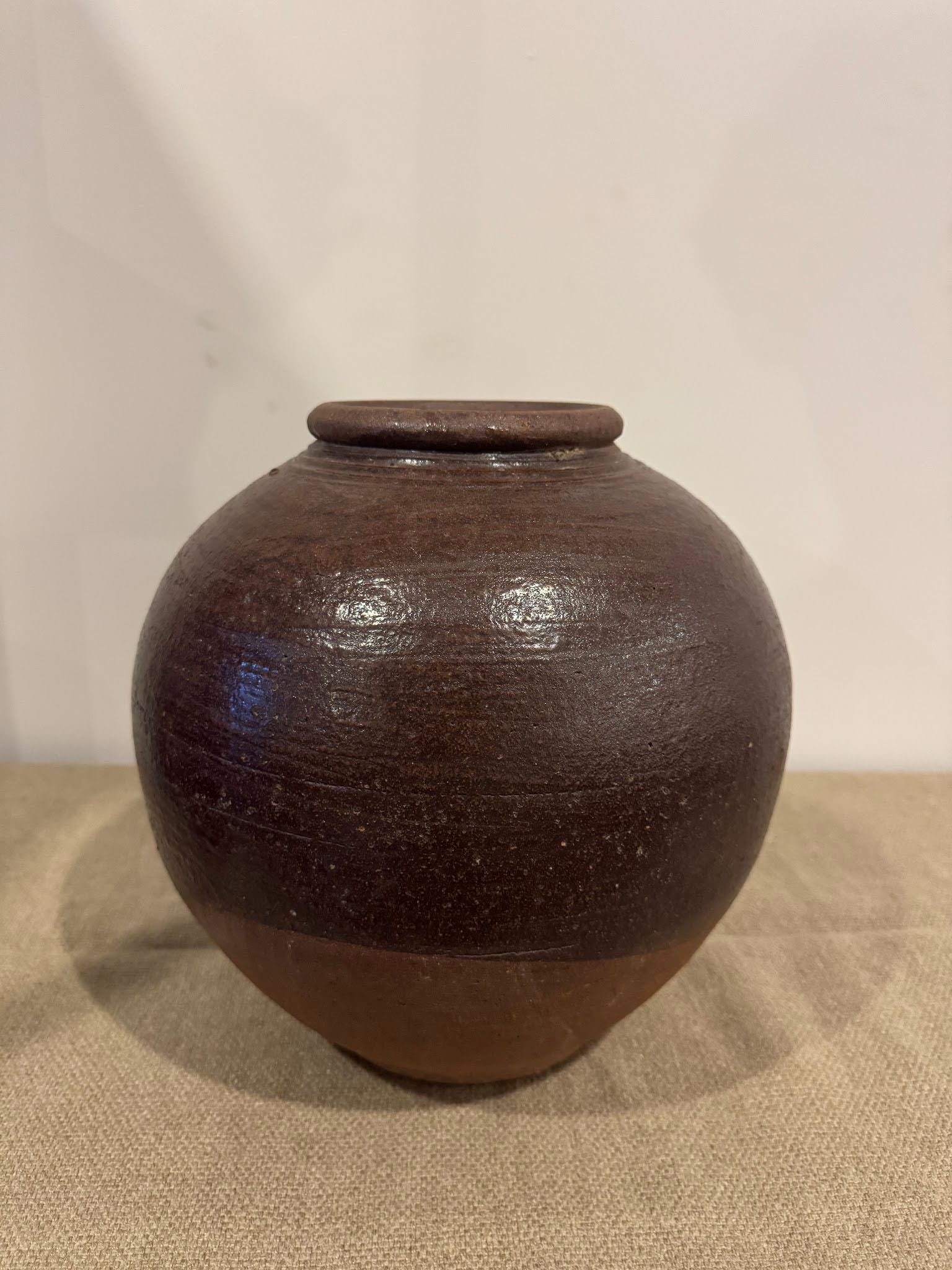 The Large Brown Pot is a round ceramic piece with a smooth, slightly glossy finish and small lid, measuring 11 inches wide. It sits on beige fabric against a light wall—an ideal statement for any setting.