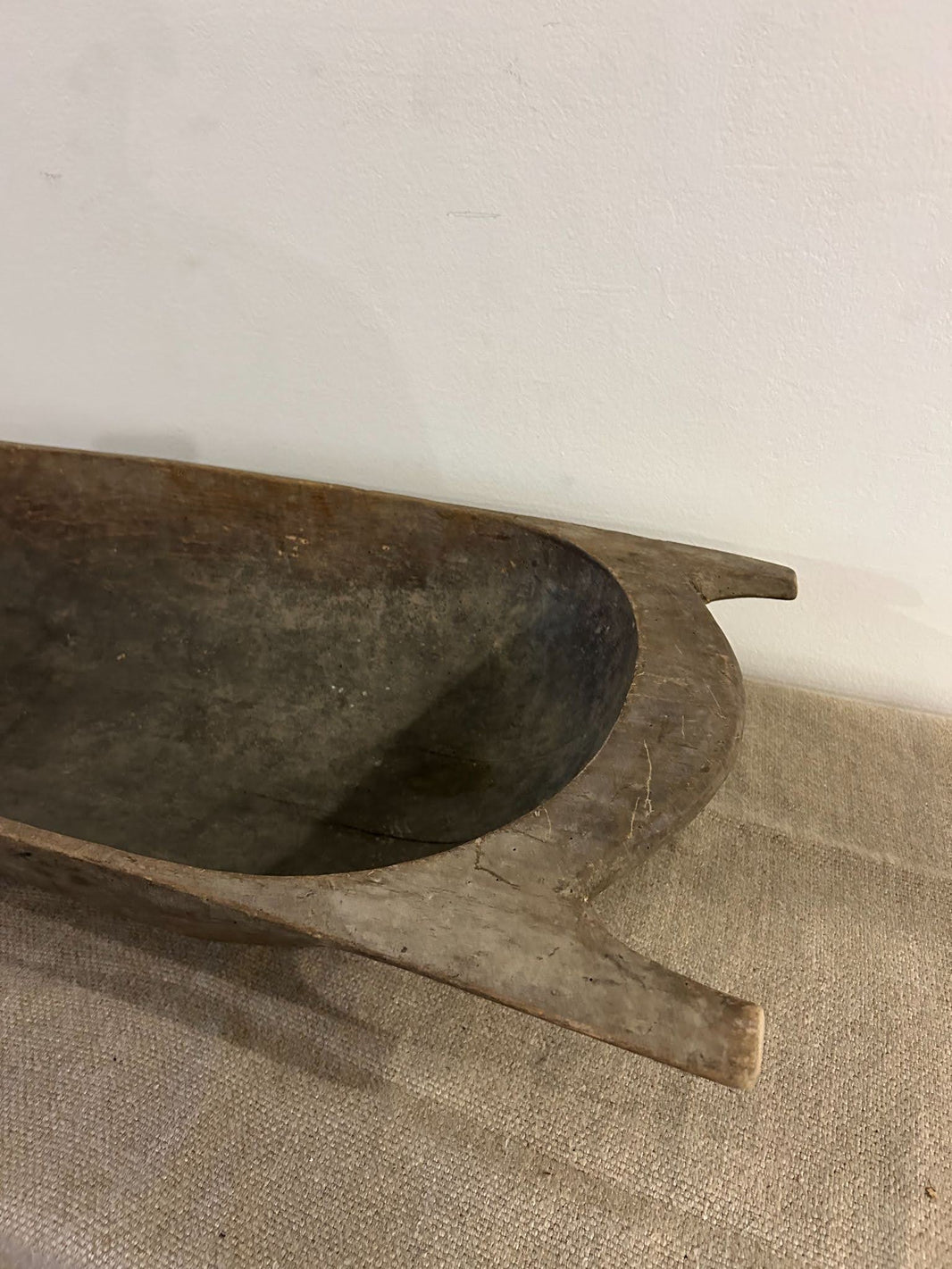 A close-up of the Large Dark Display Bowl, a rustic wooden piece with carved side handles and a smooth, weathered finish, resting on textured beige pet bed fabric against a plain white wall.