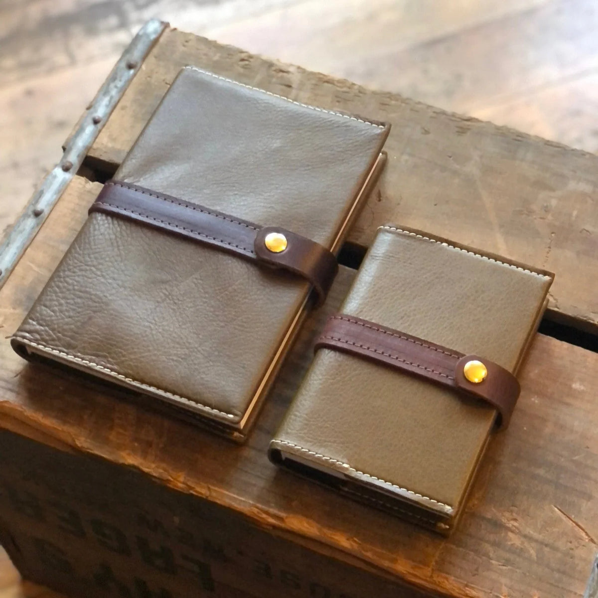 Two brown handcrafted Writer's Journals with dark straps and gold buttons rest on a rustic wooden surface. One notebook is larger than the other, making this pair ideal as a custom branded keepsake for corporate gifting.