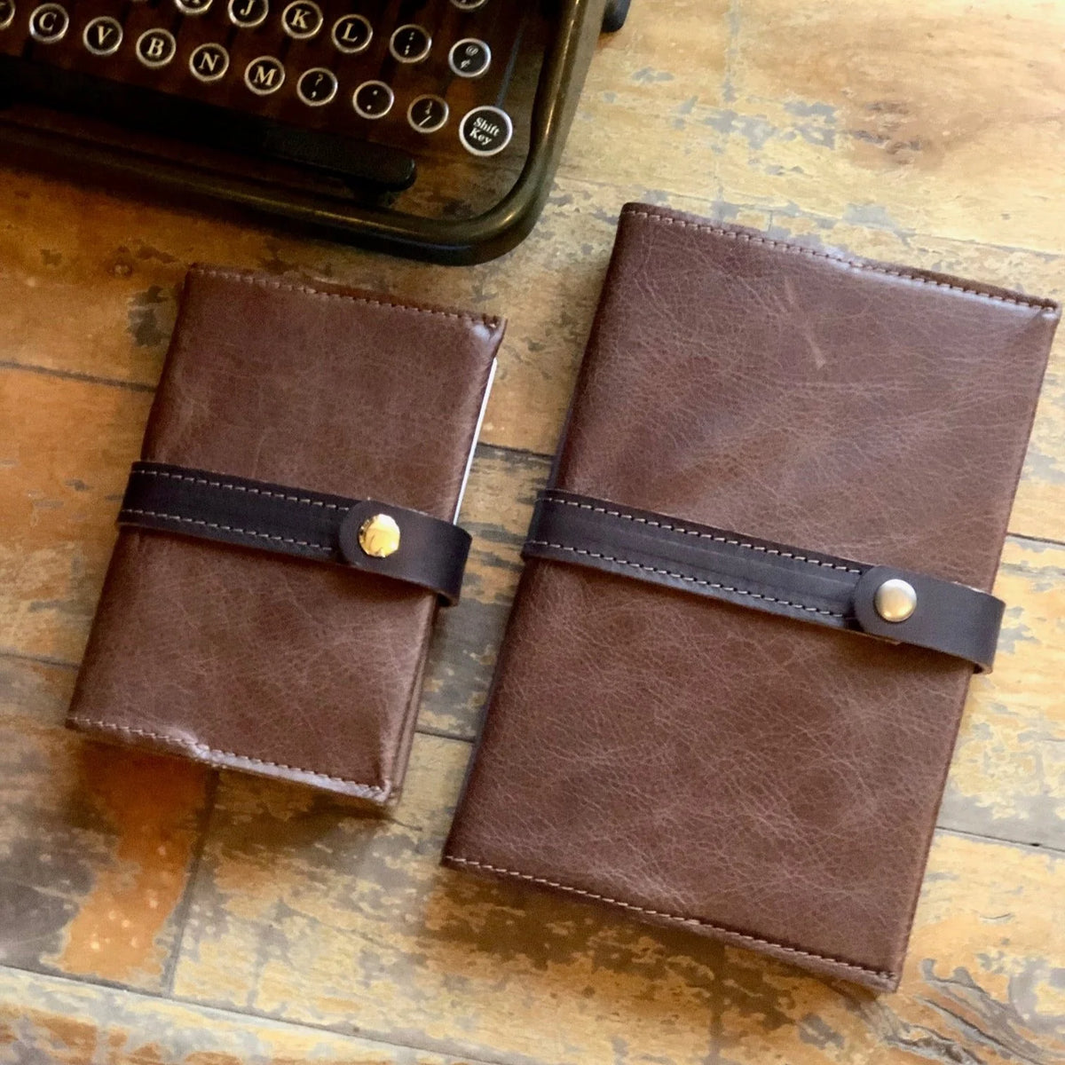 Two brown handcrafted Writer’s Journals, featuring dark straps and button closures, rest on a wooden surface beside part of a vintage typewriter—perfect as a custom branded keepsake for corporate gifting.