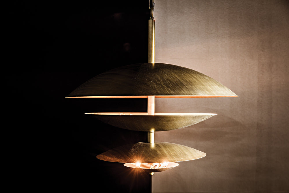 The Chandelier Two Tiered Metal with Aged Brass Finish features layered discs and artisan craftsmanship, casting a warm glow against dark or neutral backgrounds.