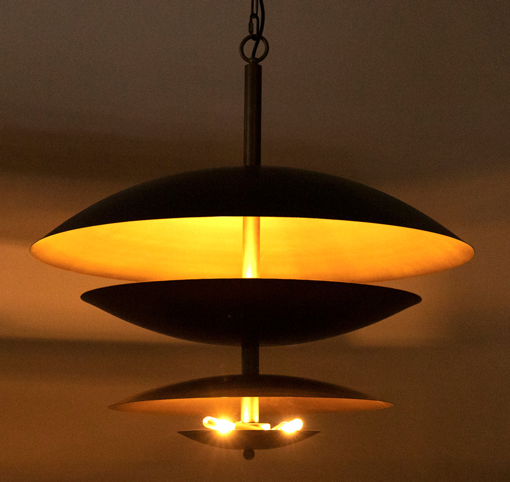 The Chandelier Two Tiered Metal with Aged Brass Finish features a modern design and emits a warm, yellow glow, creating soft ambient lighting against a neutral wall.