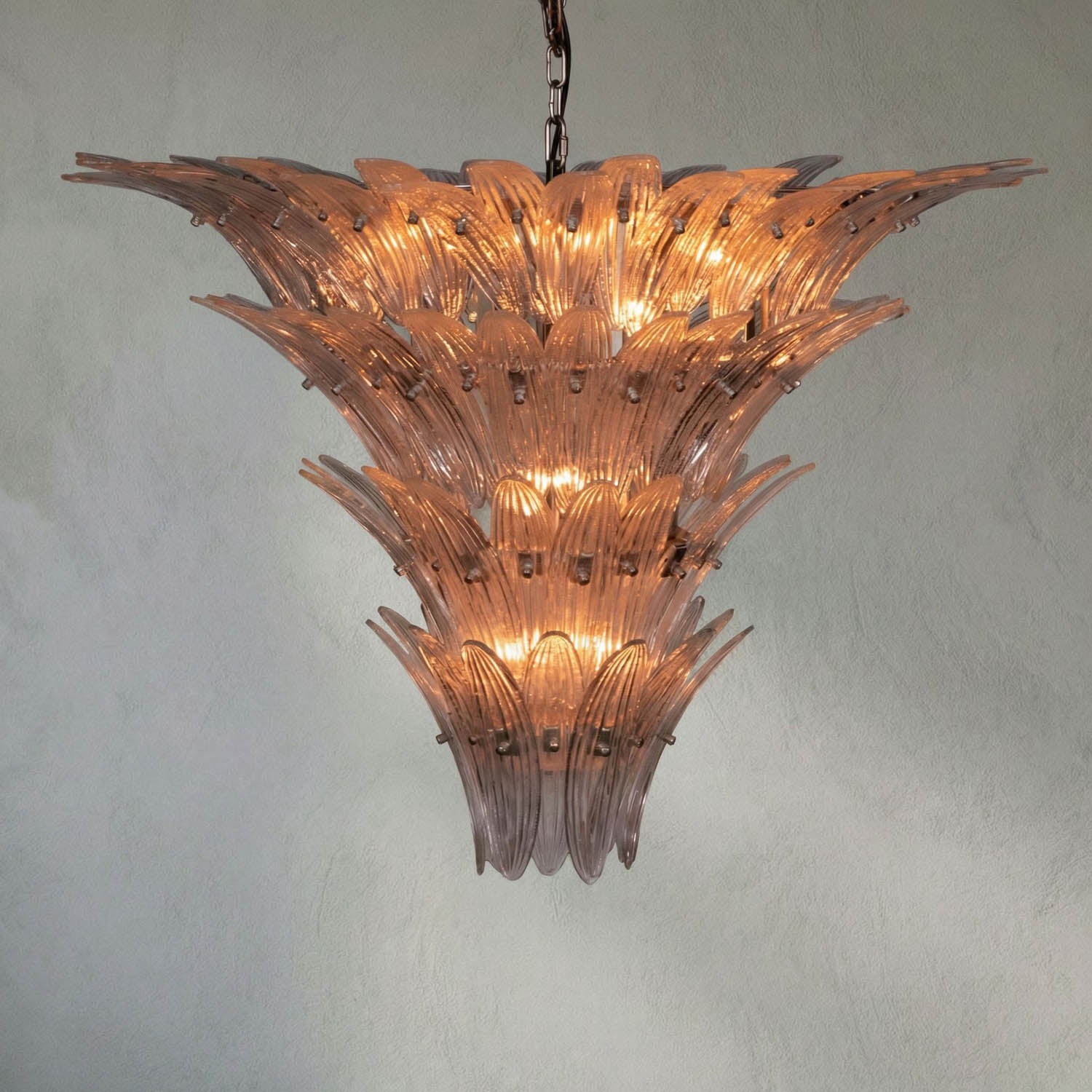 The Textured Glass Chandelier features layered, leaf-shaped textured glass pieces and multiple glowing lights, creating a luxurious statement as it hangs from the ceiling against a plain, light-colored wall.