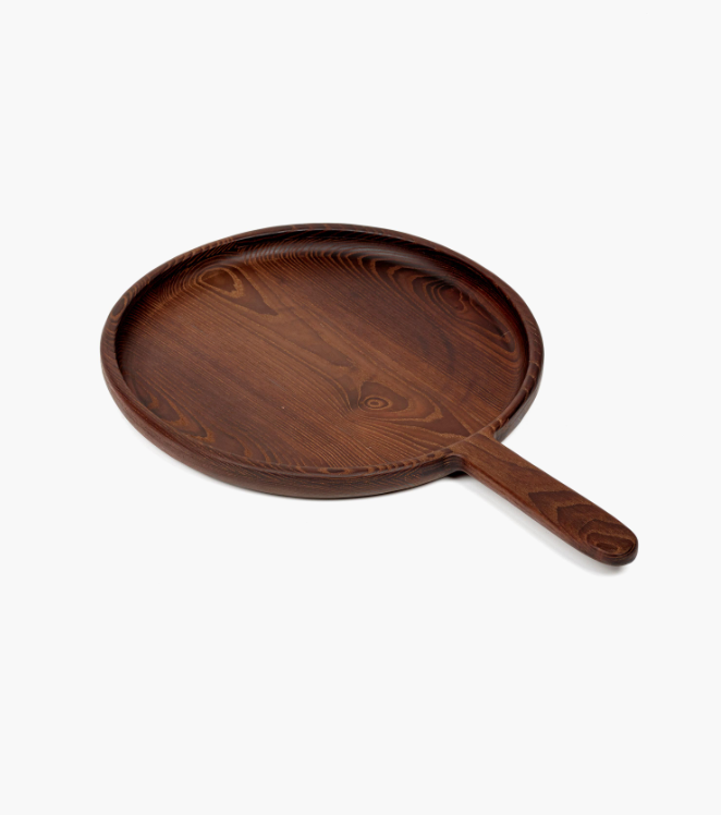 The Small Paddle Round Serving Dish is a round, dark brown platter crafted from carbonised ash with a raised edge and handle, displayed on a white background.