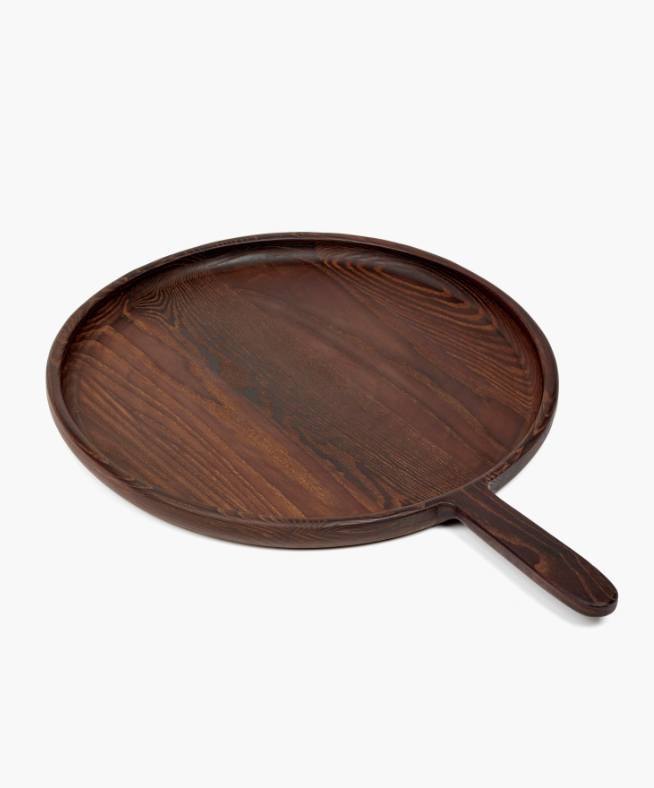 The Large Paddle Round Serving Dish is a round, dark brown wooden tray with a short handle and visible wood grain in a carbonised ash finish, measuring 19.13"L x 13.78"W x 0.98"H, shown on a white background.