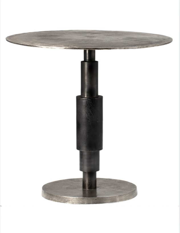 The Raw Nickel Bistro Table 30" is a round metal table with a flat top and base, connected by a thick cylindrical support. Its minimalist, industrial design is crafted from cast aluminum with a bold black patina finish.