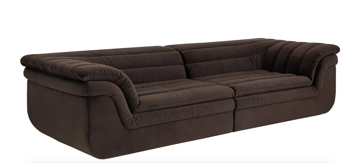 The Low-slung sculptural sofa features a modern design with dark brown velvet upholstery, channel tufting, plush curved armrests, and deep cushioned seats, shown against a plain white background.