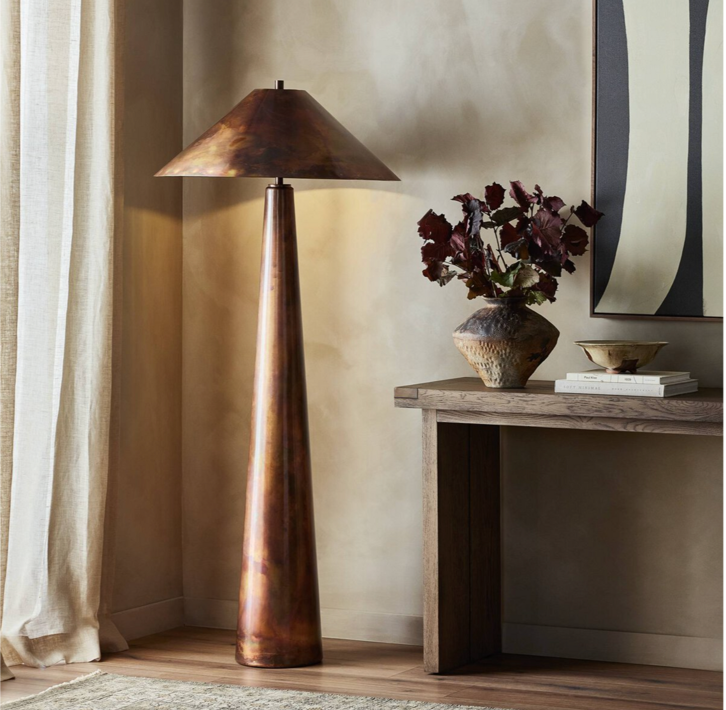 The Romani Floor Lamp, with its tall, cone-shaped design and gleaming iridescent finish, stands beside a wooden console table topped with a vase of dark red leaves, decorative bowl, and books, set against a softly lit wall featuring modern art.
