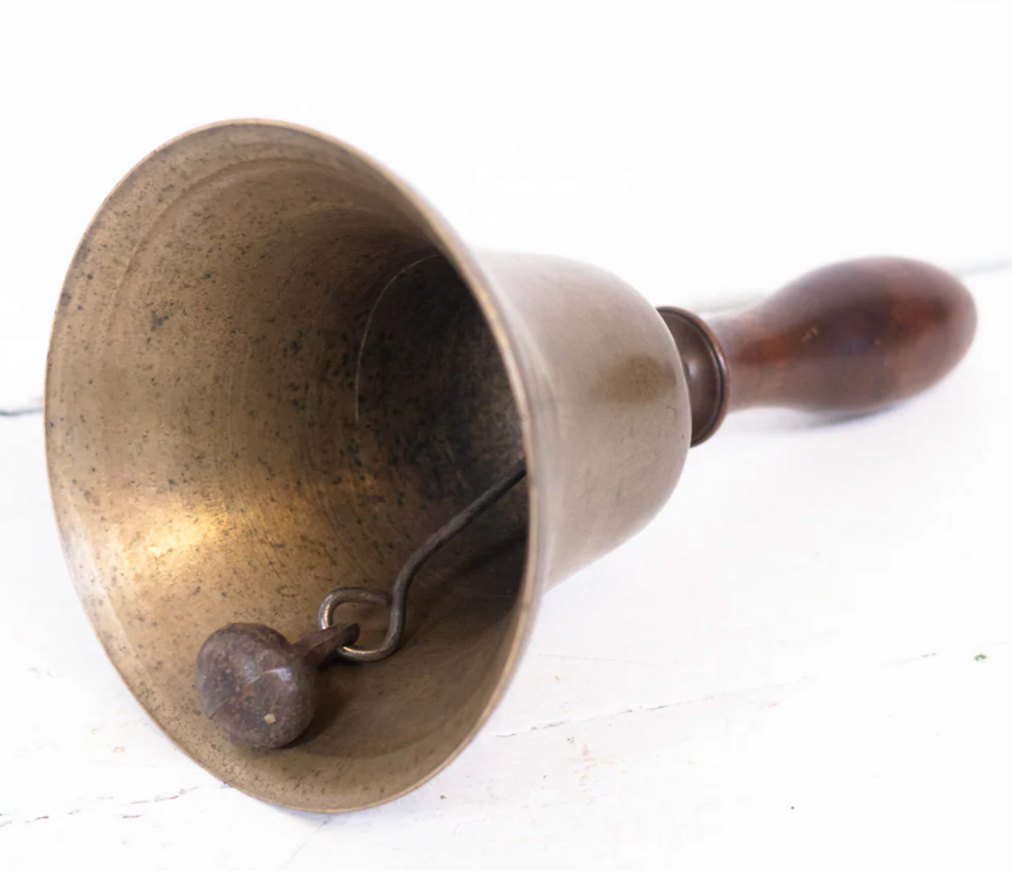 A Brass Bell with a wooden handle is displayed on its side on a white surface, revealing the clapper inside.